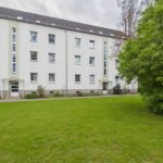 Mehrfamilienhaus mit weißen Wänden und grüner Rasenfläche im Vordergrund, umgeben von Bäumen.