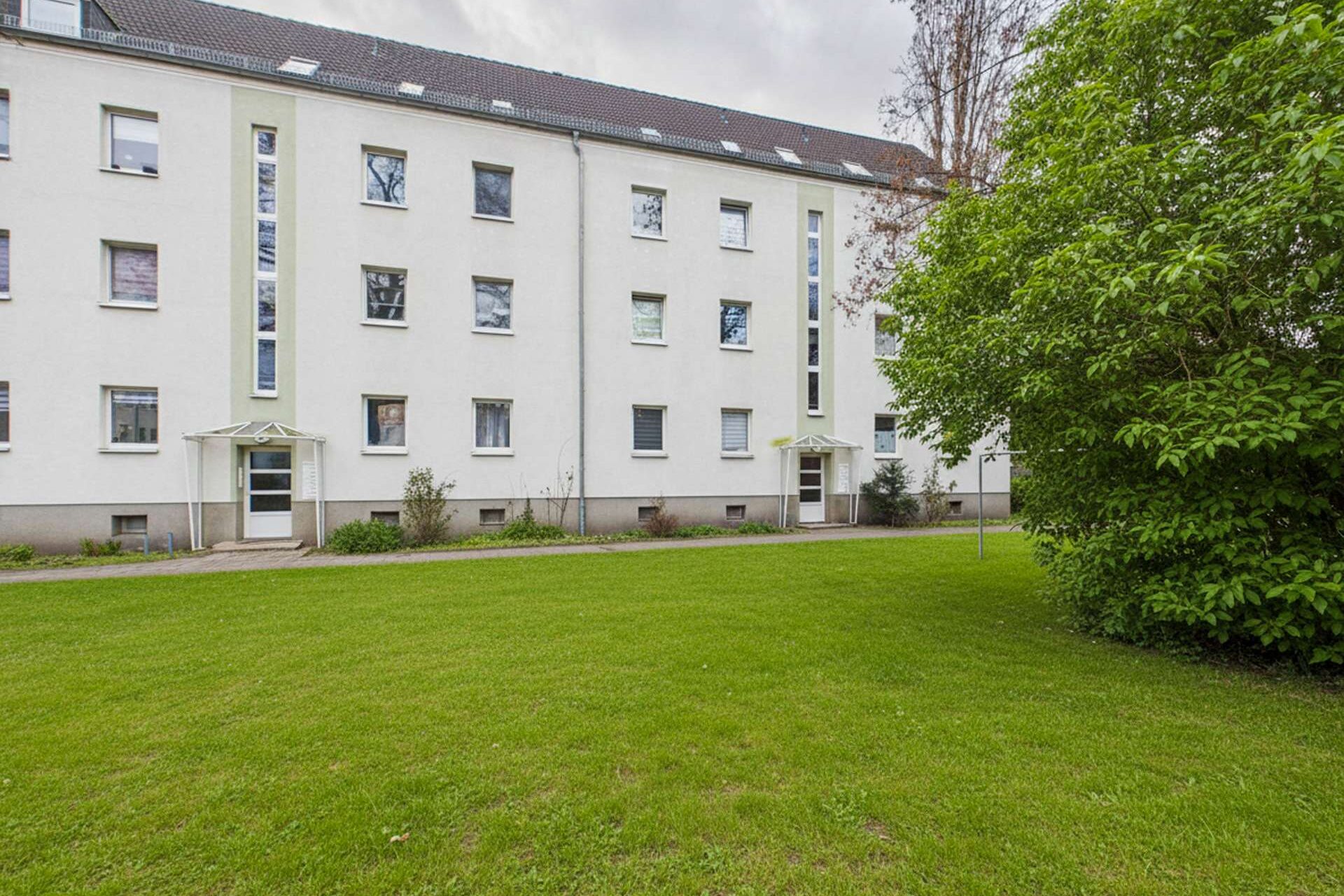 Mehrfamilienhaus mit weißen Wänden und grüner Rasenfläche im Vordergrund, umgeben von Bäumen.