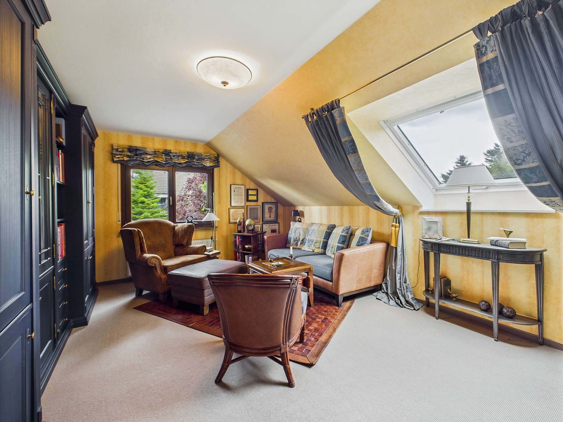 Gemütliches Dachzimmer mit einem Sofa, Sessel, Bücherregal und einem Fenster, das Licht hereinlässt.