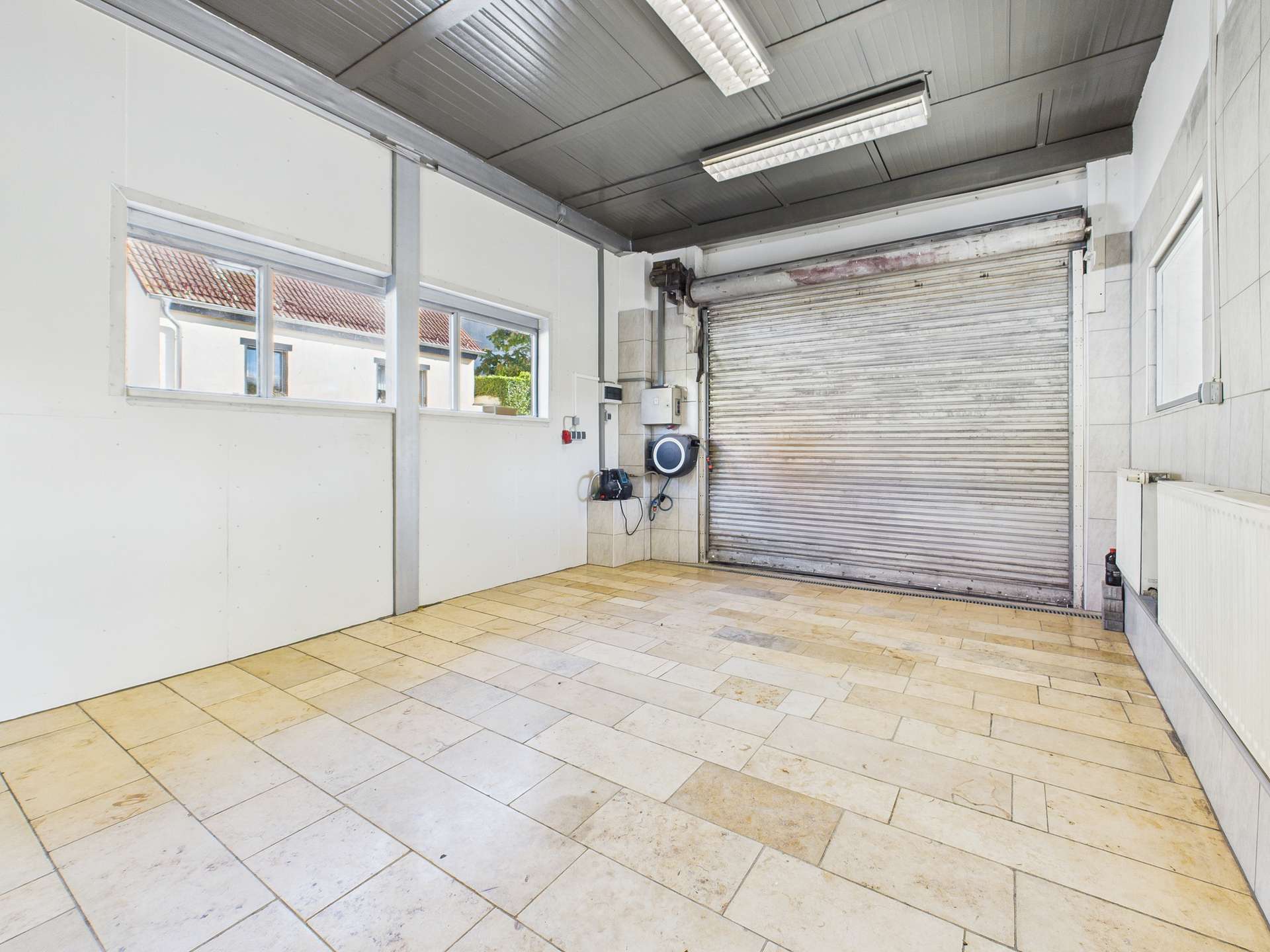 Leerer Raum mit Fliesenboden, Fenster und einem rollbaren Garagentor im Hintergrund.