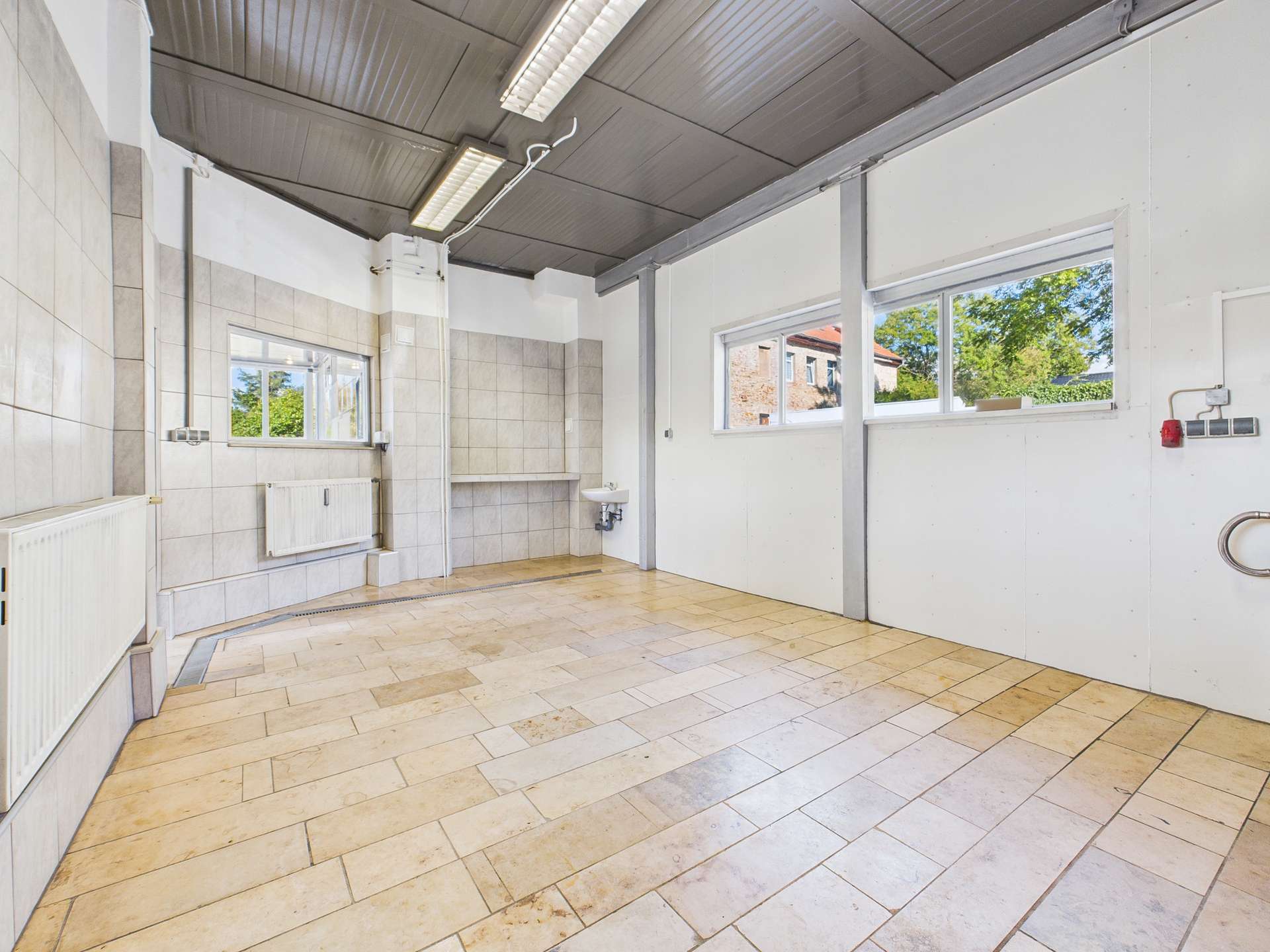 Heller, leerer Raum mit Fliesenboden, Fenstern und einem Waschbecken an der Wand.