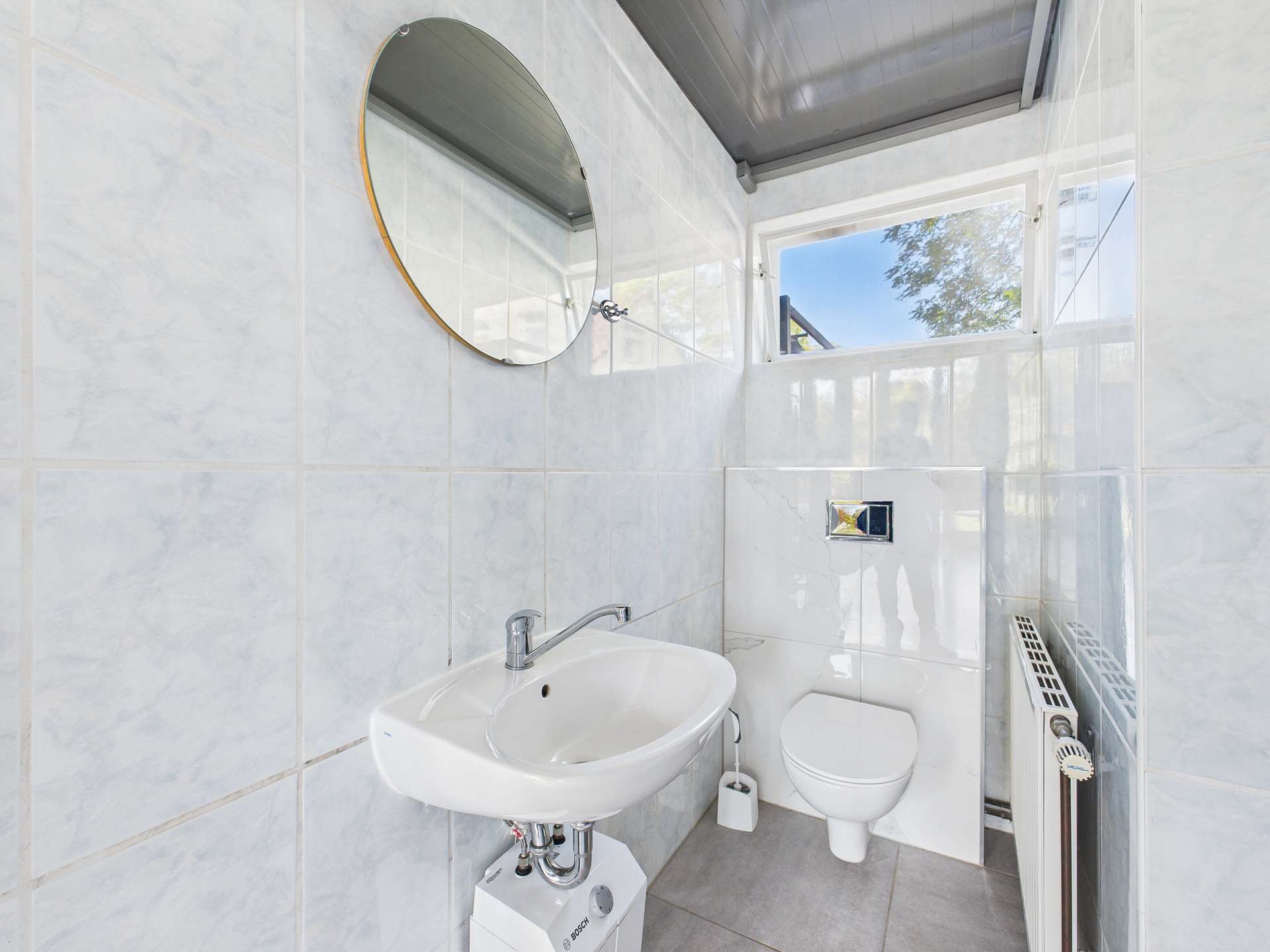Badezimmer mit Waschbecken, WC und runder Wandspiegel, gefliest mit hellen, modernen Fliesen und Fensterlicht.