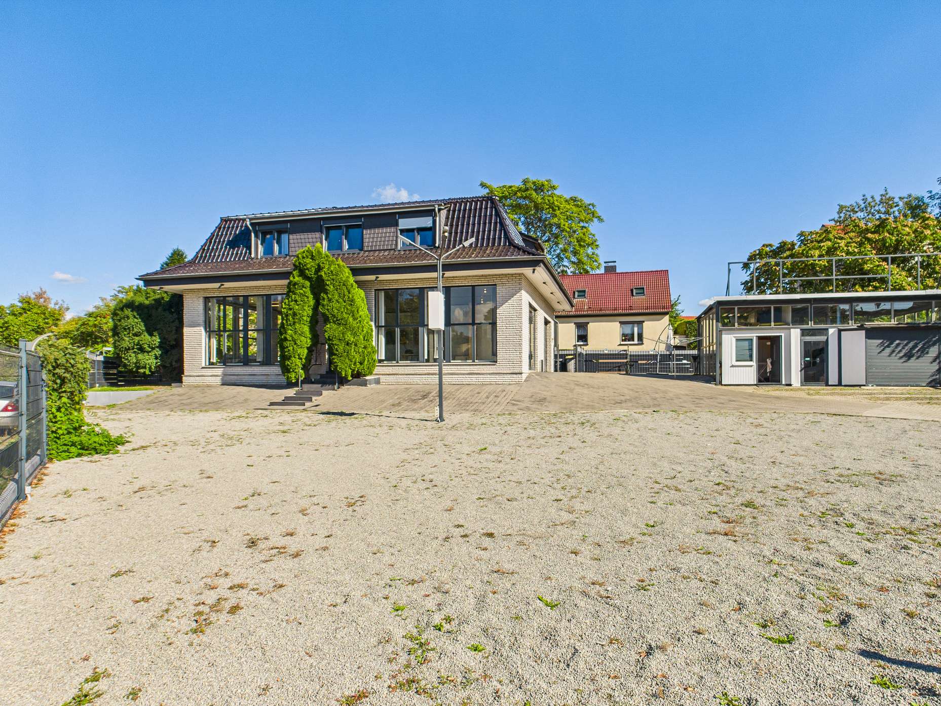 Modernes Haus mit großen Fenstern und gepflegtem Garten, umgeben von einem Kiesplatz und Bäumen.