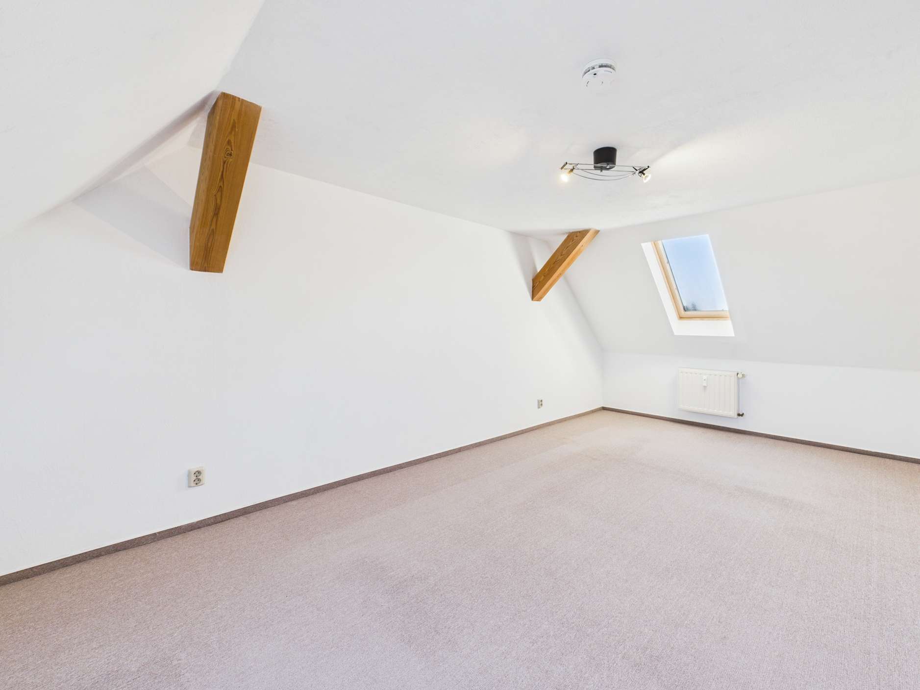 Leerer Dachraum mit schrägen Wänden, Holzträgern und einem kleinen Fenster, das Tageslicht einlässt.