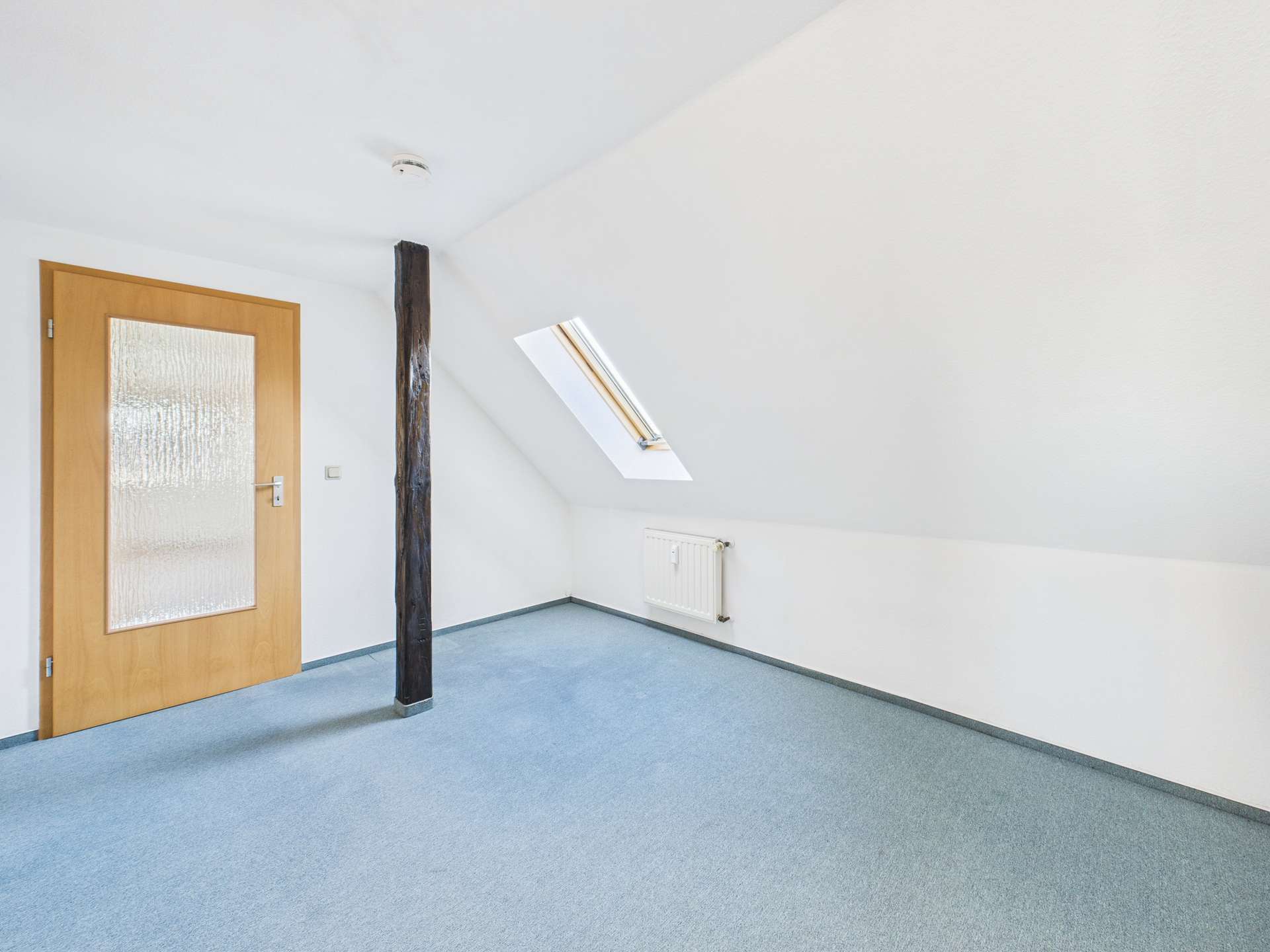 Heller Dachraum mit blauer Teppichfläche, Holzsäule und Fenster, das Tageslicht hereinlässt.