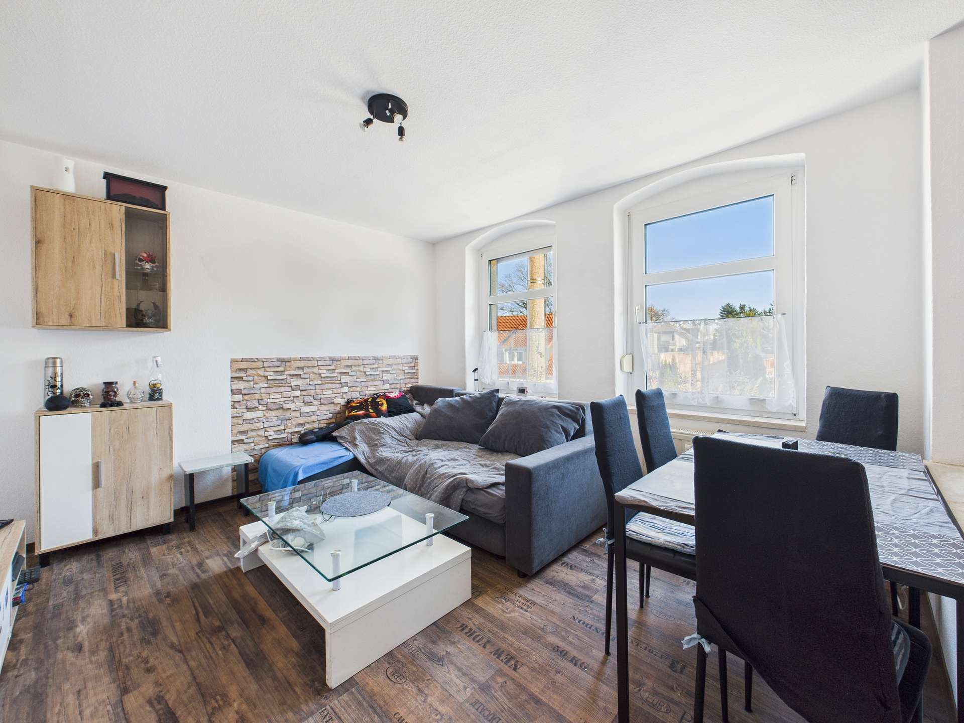 Helle Wohnküche mit einem grauen Sofa, einem Glastisch und Holzregalen, großen Fenstern und moderner Einrichtung.