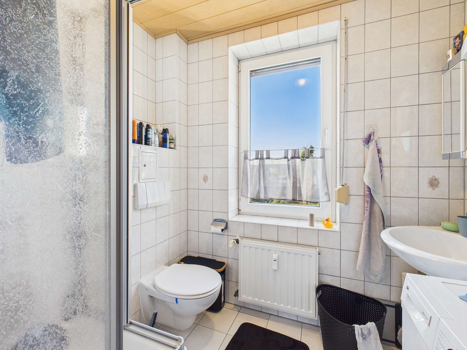 Kleines Badezimmer mit Dusche, WC, Waschbecken und Fenster, gefliest mit hellen Fliesen und dekorativen Elementen.