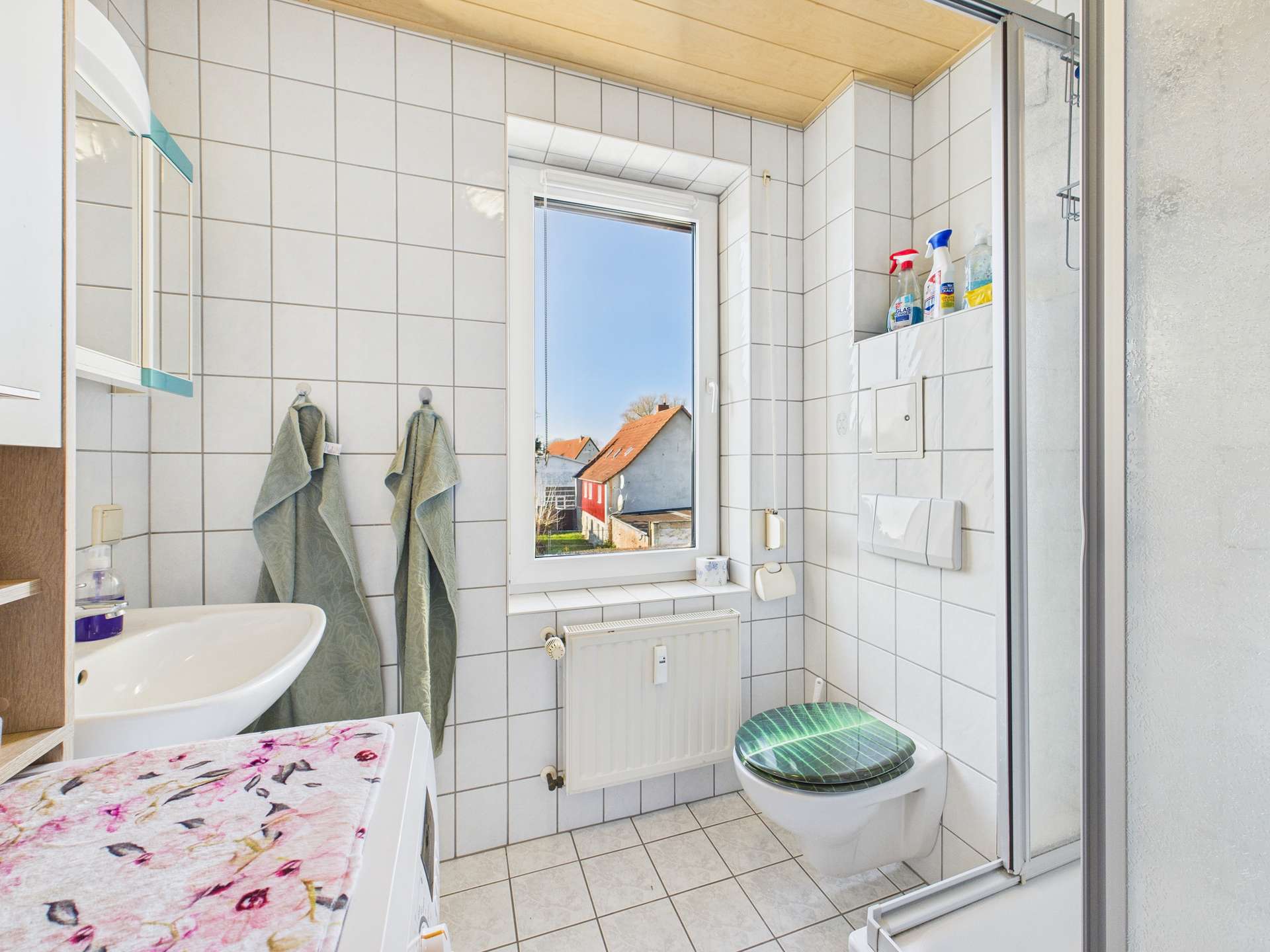 Badezimmer mit Fenster, Waschbecken, WC und gefliesten Wänden; Handtücher und Pflegeprodukte sind sichtbar.