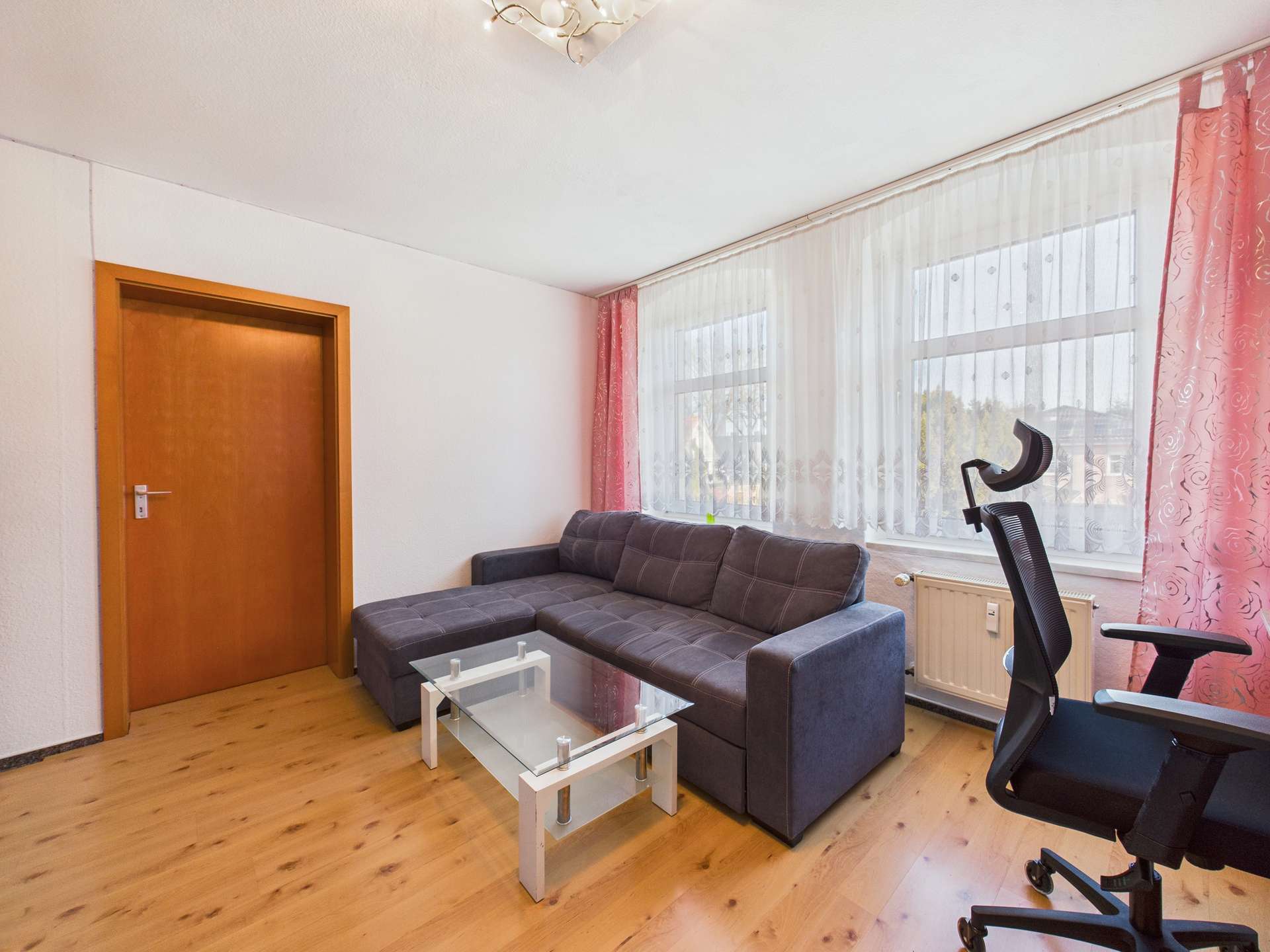 Helles Wohnzimmer mit grauem Sofa, Glastisch, Schreibtischstuhl und großen Fenstern mit Vorhängen.