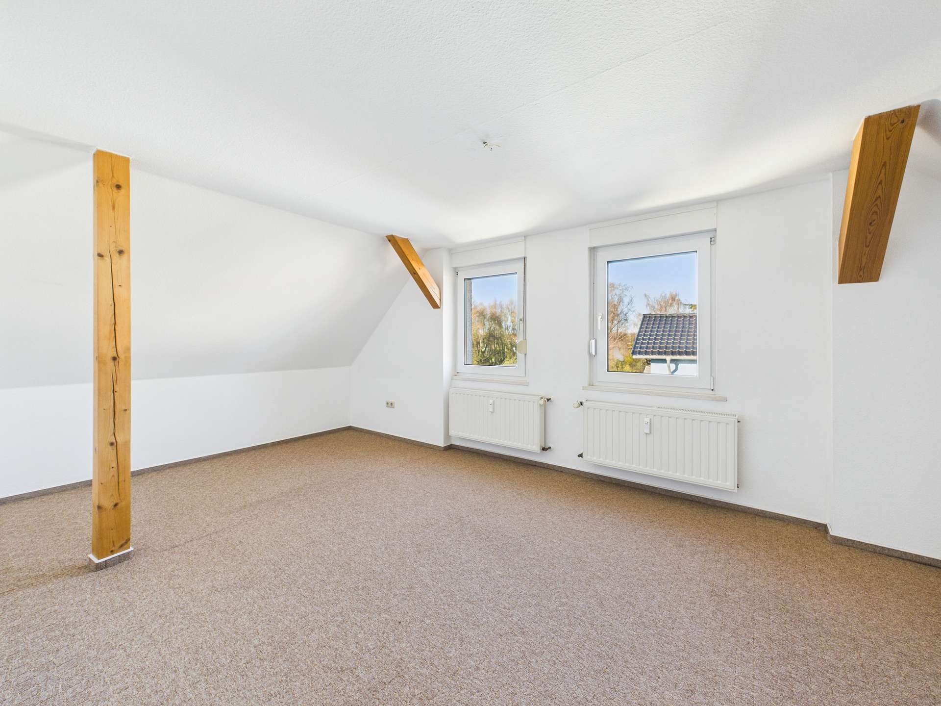 Heller, leerer Dachraum mit Holzstützen und zwei Fenstern, Teppichboden und Heizkörpern an den Wänden.