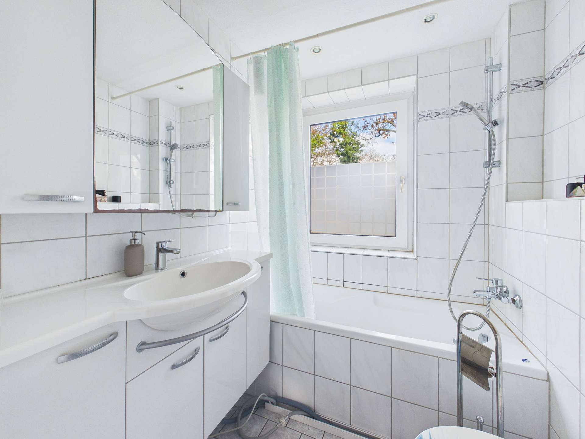 Helles Badezimmer mit Badewanne, Waschbecken, Spiegel und Fenster, gefliest in Weiß und mit grüner Duschvorhang.