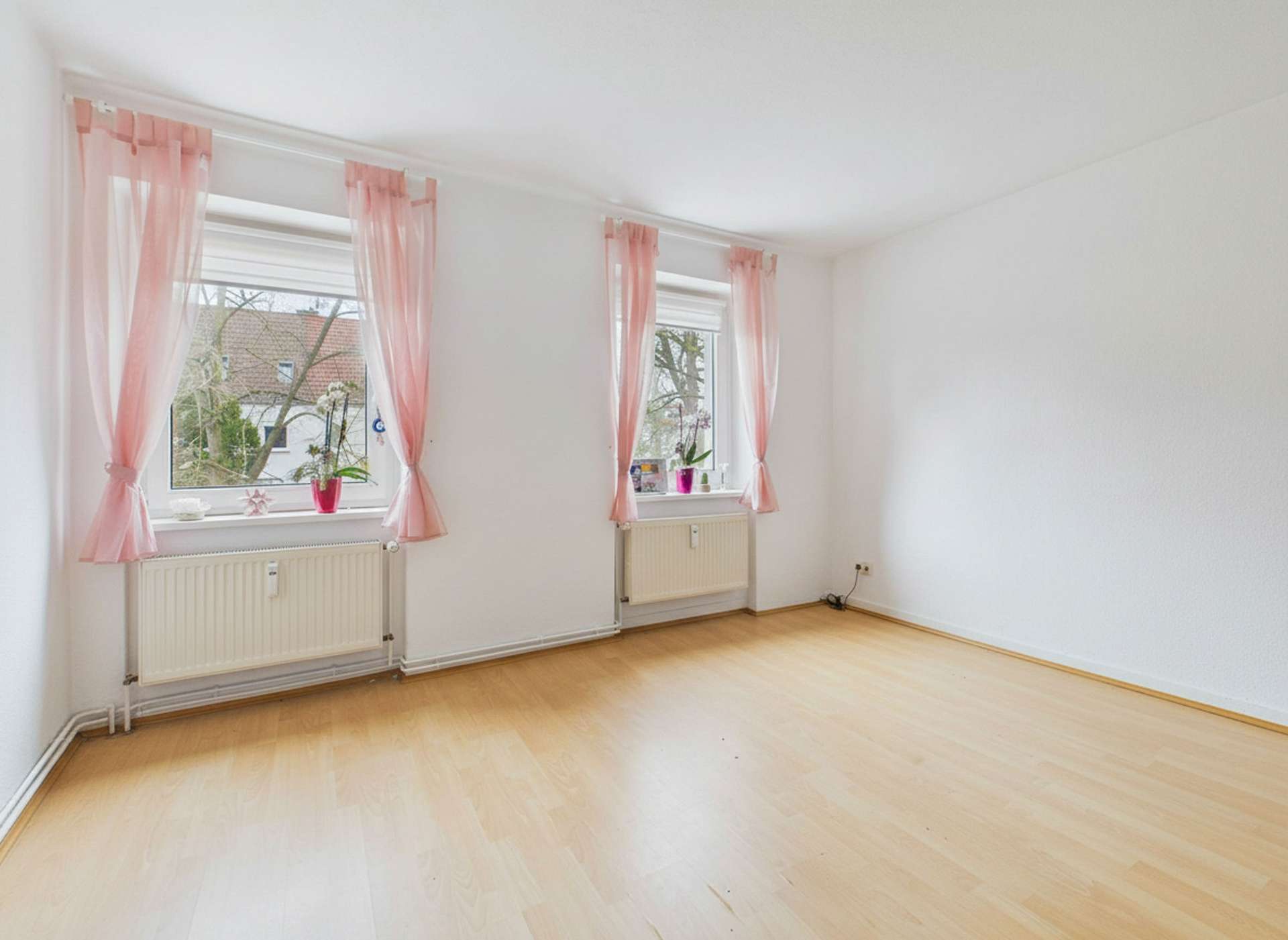 Helles, leeres Zimmer mit zwei Fenstern und rosa Vorhängen, Holzfußboden und neutralen Wänden.