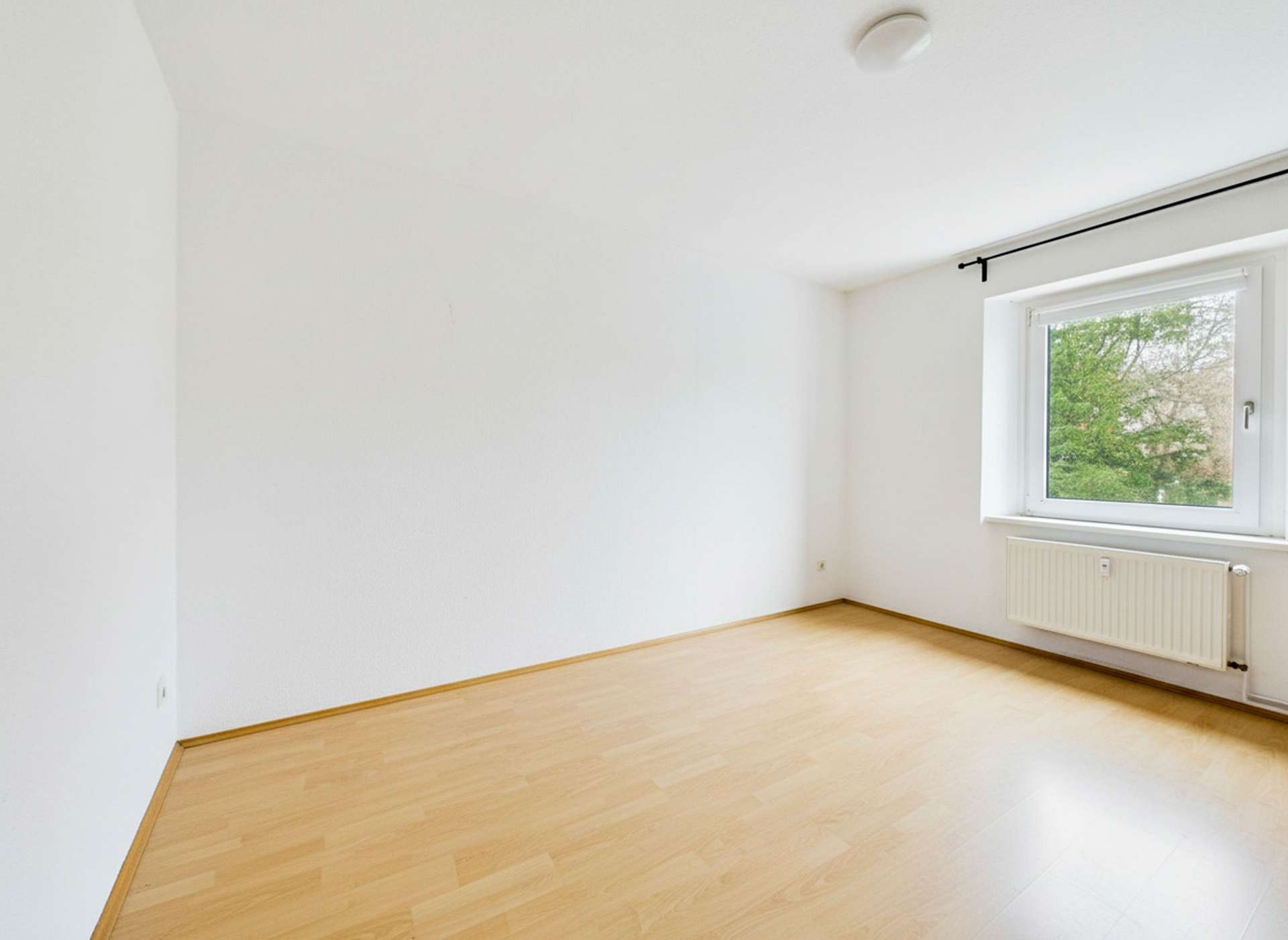 Heller, leerer Raum mit Parkettboden und Fenster, das auf einen grünen Außenbereich blickt.