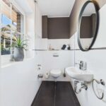 Modernes Badezimmer mit WC, Waschbecken, runder Spiegelfläche und Pflanzenarrangement. Helle Fliesen und dunkler Boden.