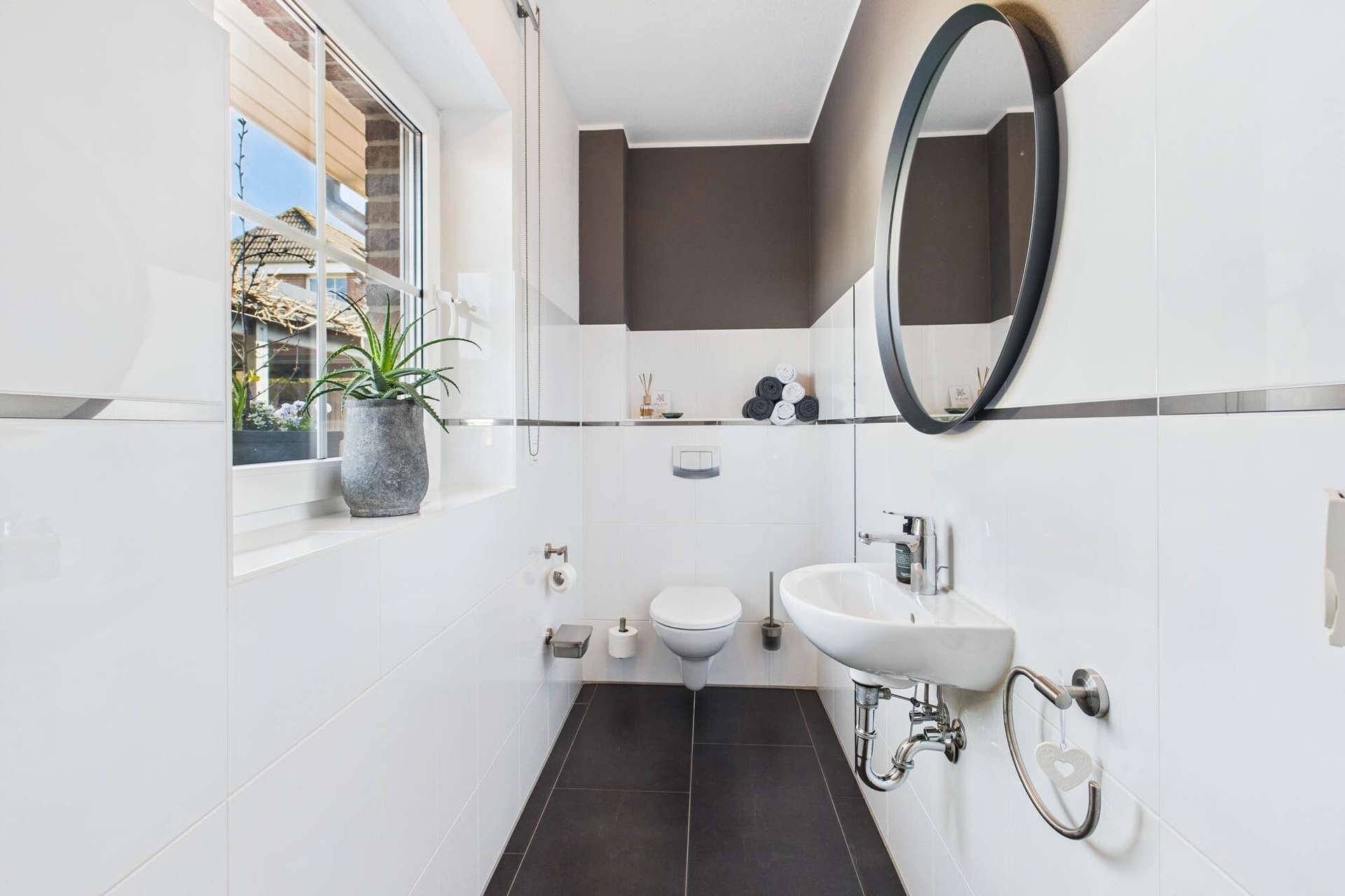 Modernes Badezimmer mit WC, Waschbecken, runder Spiegelfläche und Pflanzenarrangement. Helle Fliesen und dunkler Boden.