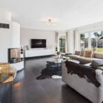 Modernes Wohnzimmer mit beiger Couch, Kamin, TV und großen Fenstern, die auf einen Garten blicken. Elegante Dekorationselemente.