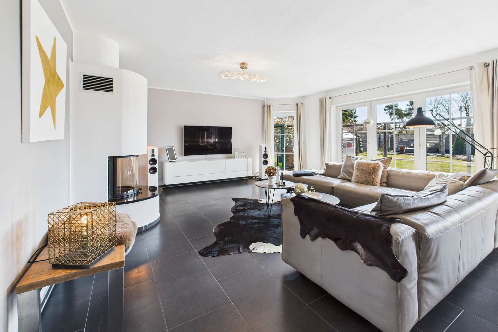 Modernes Wohnzimmer mit beiger Couch, Kamin, TV und großen Fenstern, die auf einen Garten blicken. Elegante Dekorationselemente.