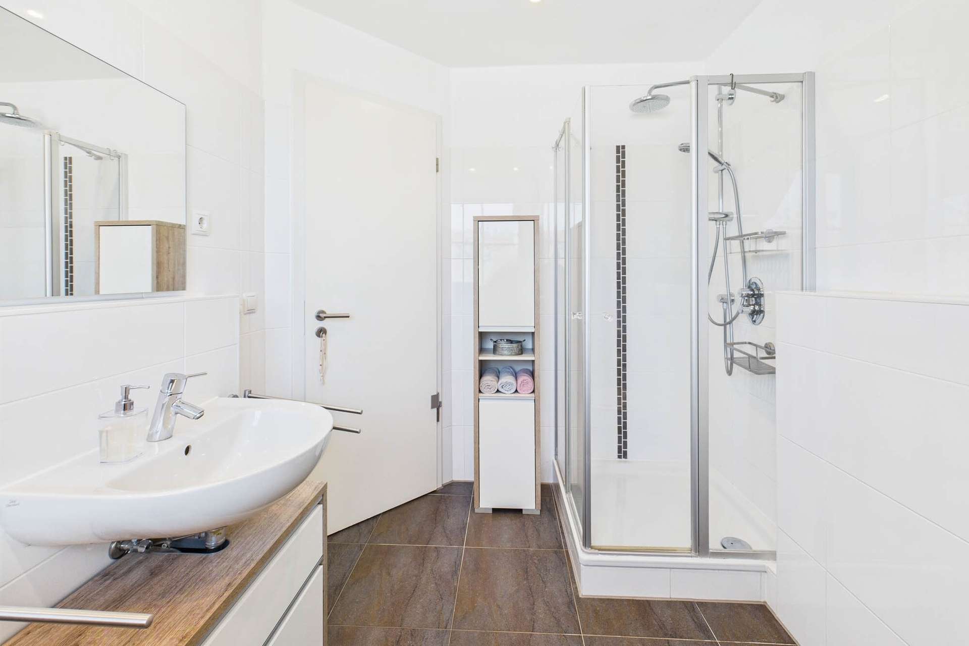 Modernes Badezimmer mit einer Dusche, Waschbecken und einem schmalen Schrank für Handtücher und Pflegeprodukte.