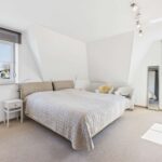 Helles Schlafzimmer mit Doppelbett, Nachttisch, Sessel und großem Fenster, das Tageslicht hereinlässt.