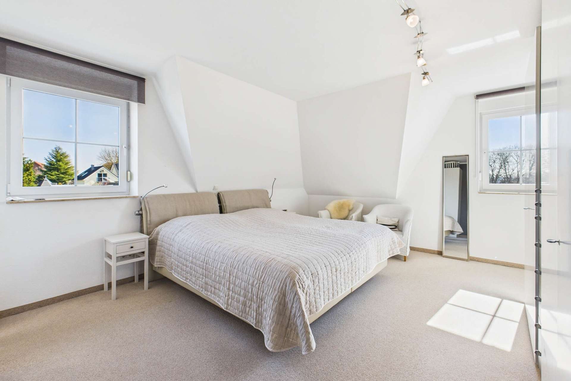 Helles Schlafzimmer mit Doppelbett, Nachttisch, Sessel und großem Fenster, das Tageslicht hereinlässt.
