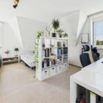 Helles Zimmer mit einem Bett, Bücherregal, Schreibtisch und Pflanzen, große Fenster mit Blick ins Freie.