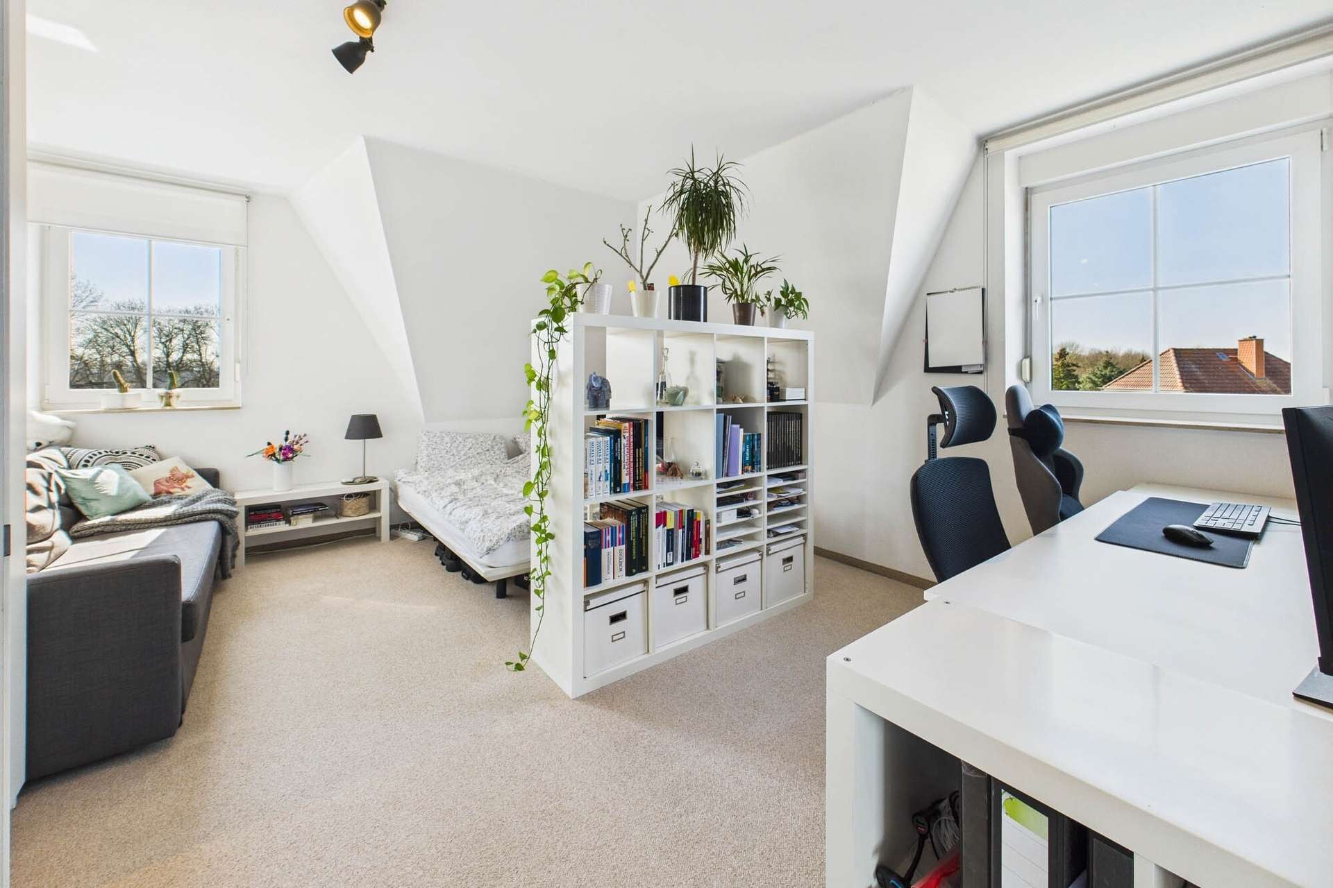 Helles Zimmer mit einem Bett, Bücherregal, Schreibtisch und Pflanzen, große Fenster mit Blick ins Freie.