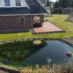 Ein gepflegter Garten mit einem Teich und einem modernen Haus im Hintergrund, umgeben von Rasen und Wegen.