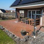 Terrasse mit roten Pflastersteinen, Holz-Tisch und Stühlen, umgeben von Pflanzen und einem gepflegten Garten.