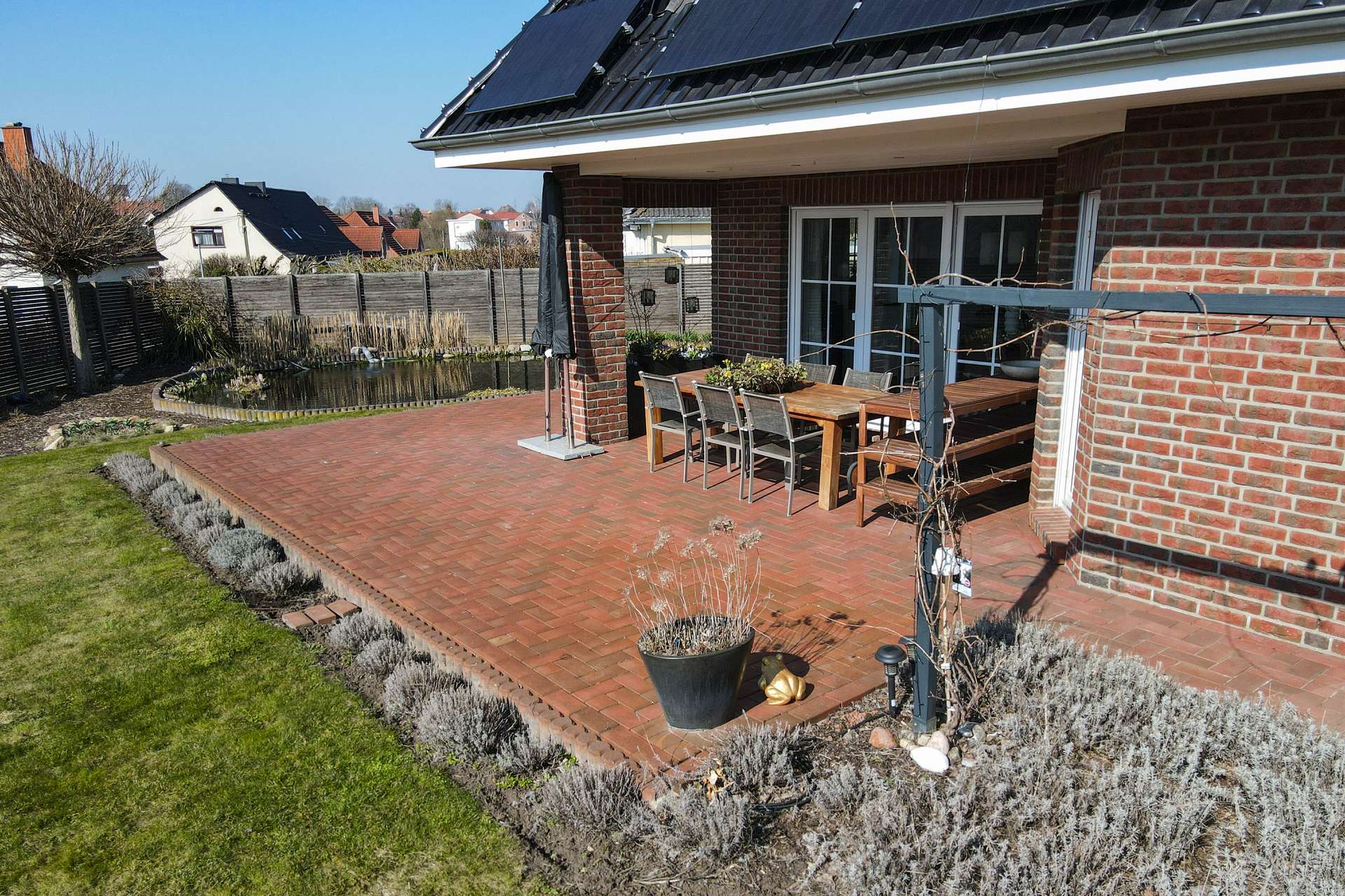 Terrasse mit roten Pflastersteinen, Holz-Tisch und Stühlen, umgeben von Pflanzen und einem gepflegten Garten.