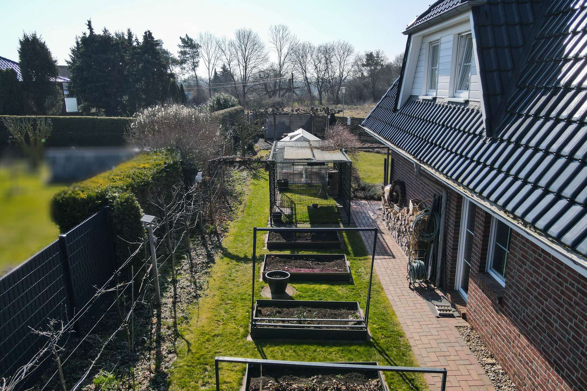 Gartenansicht mit Hochbeeten, gepflastertem Weg und einem modernen Wohnhaus im Hintergrund unter klarem Himmel.