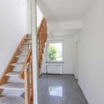 Heller Flur mit Treppe aus Holz, Fenster mit Blick ins Freie und grauem Fliesenboden.