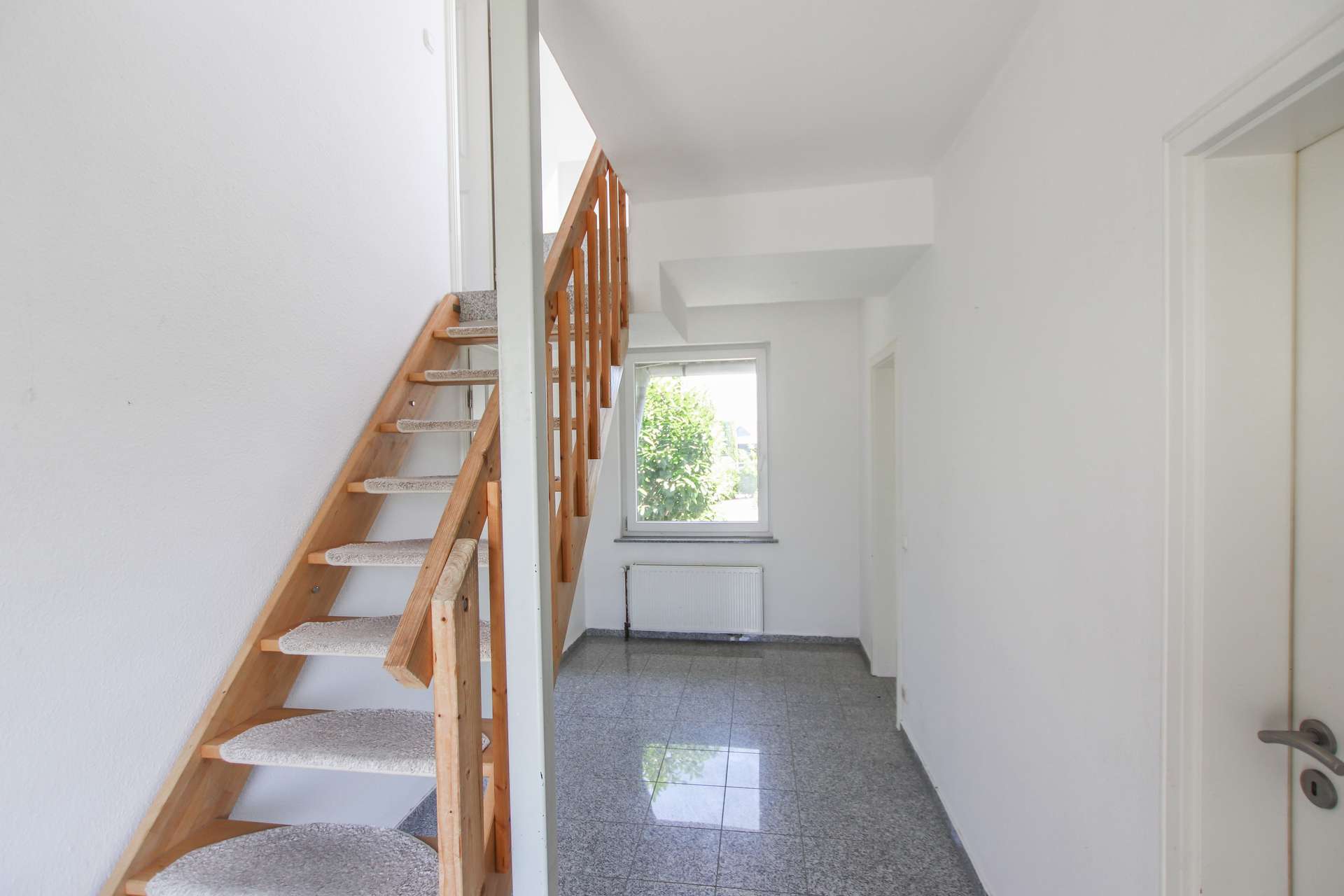Heller Flur mit Treppe aus Holz, Fenster mit Blick ins Freie und grauem Fliesenboden.