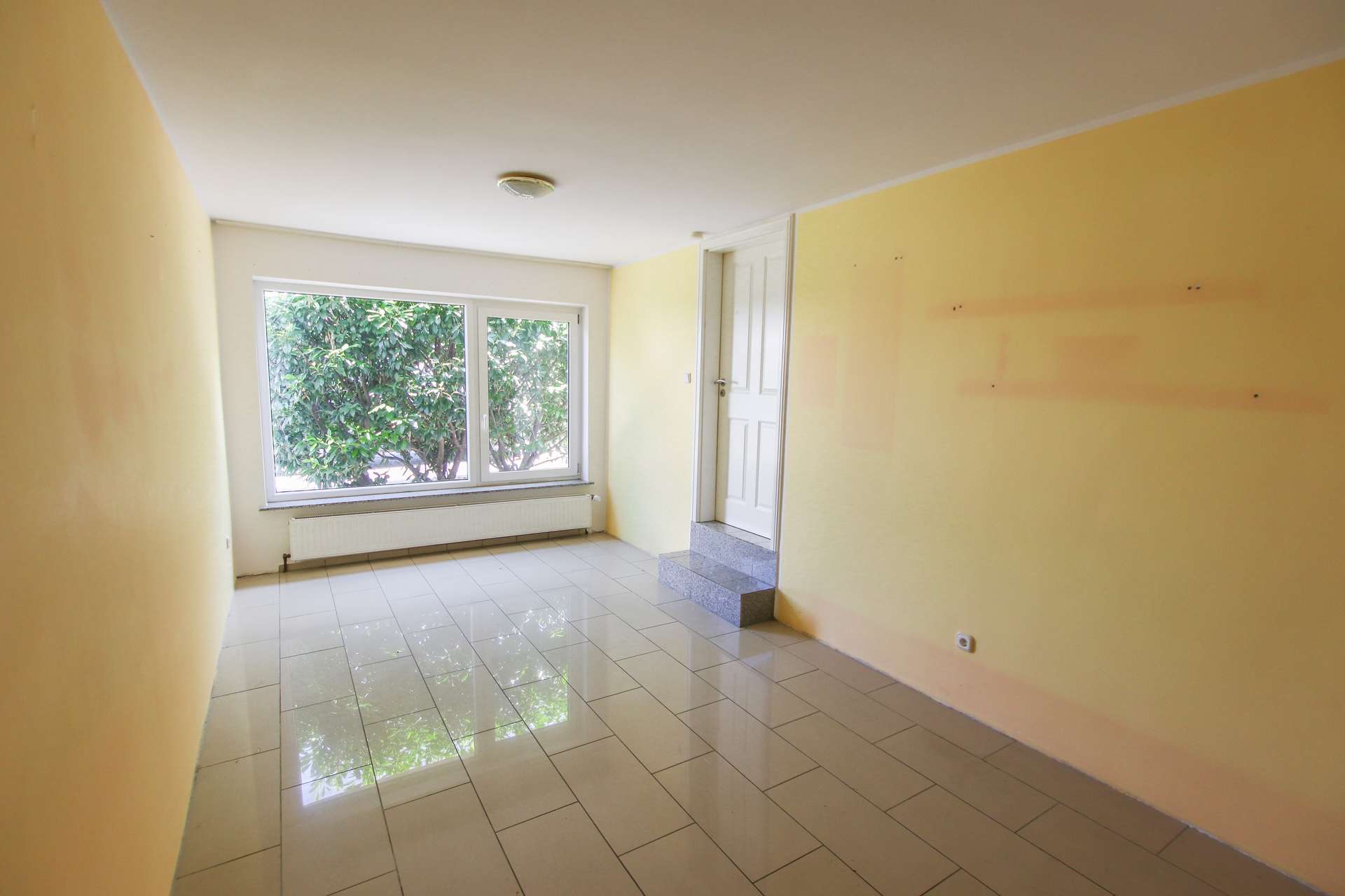 Heller Raum mit gelben Wänden, großem Fenster und Fliesenboden, ein Treppenaufgang im Hintergrund.