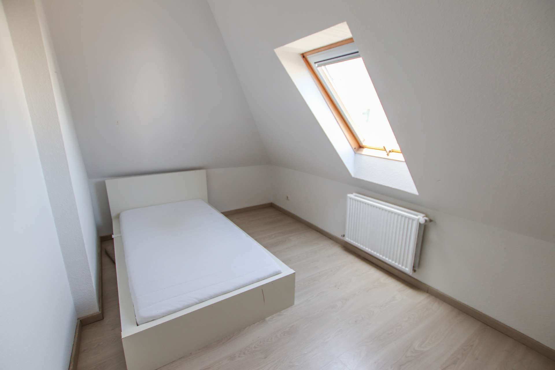 Helle Dachgeschosszimmer mit schrägen Wänden, einem Bett ohne Matratze und einem Fenster.
