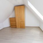 Leeres Dachzimmer mit Holzschrank und Kommode, schrägen Wänden und hellem Fußboden.