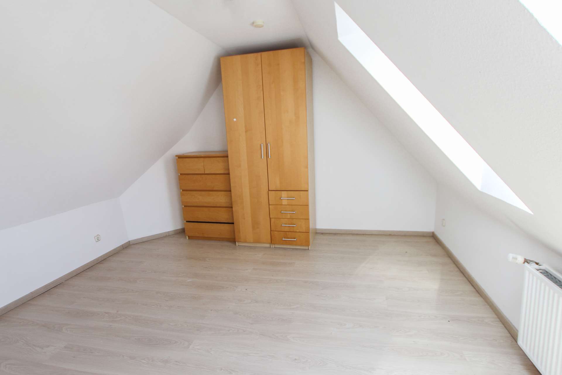 Leeres Dachzimmer mit Holzschrank und Kommode, schrägen Wänden und hellem Fußboden.