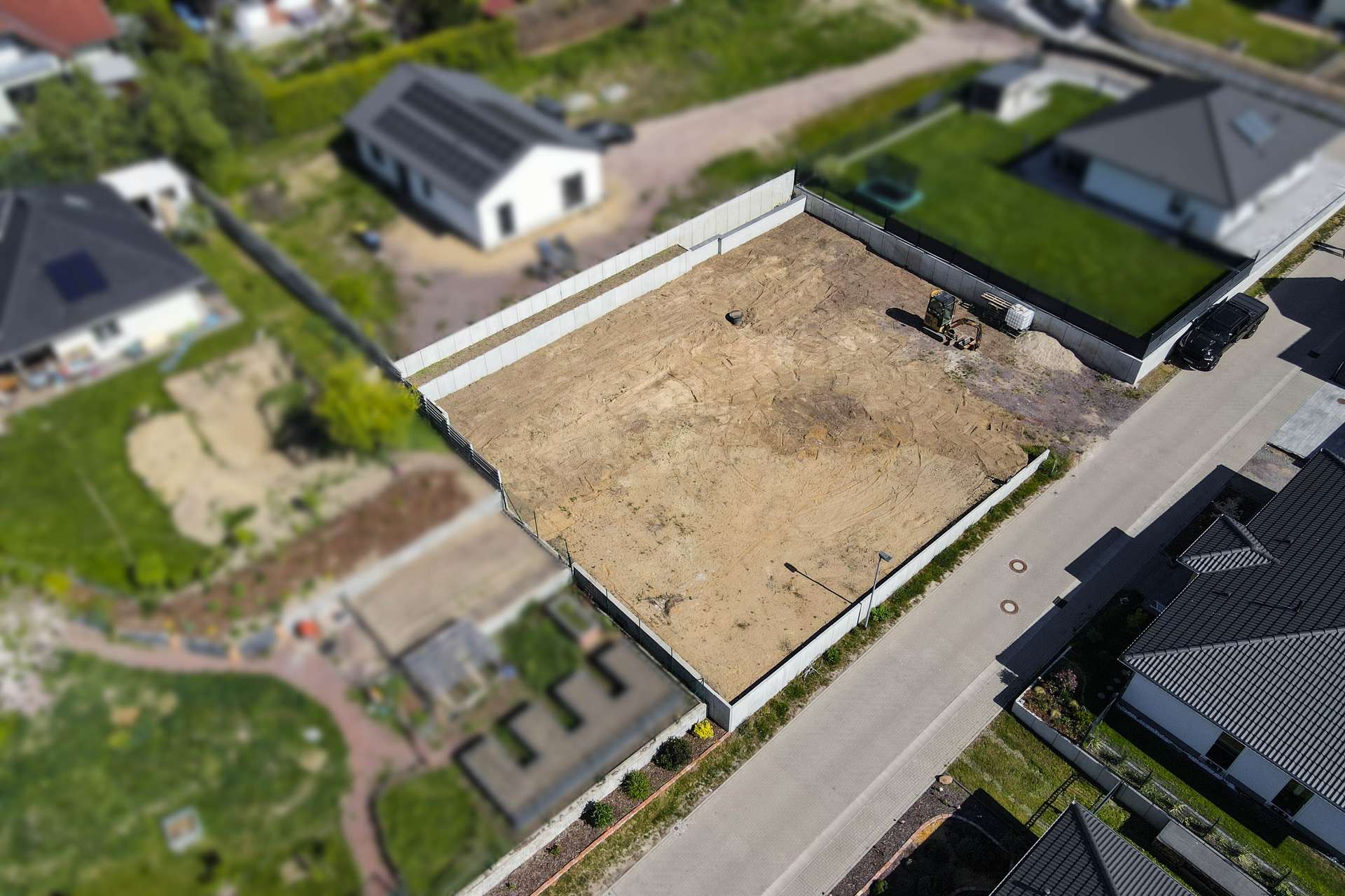 Eine leere, unbebaute Baustelle umgeben von Wohnhäusern, mit einem gepflasterten Weg und grünem Gartenbereich.