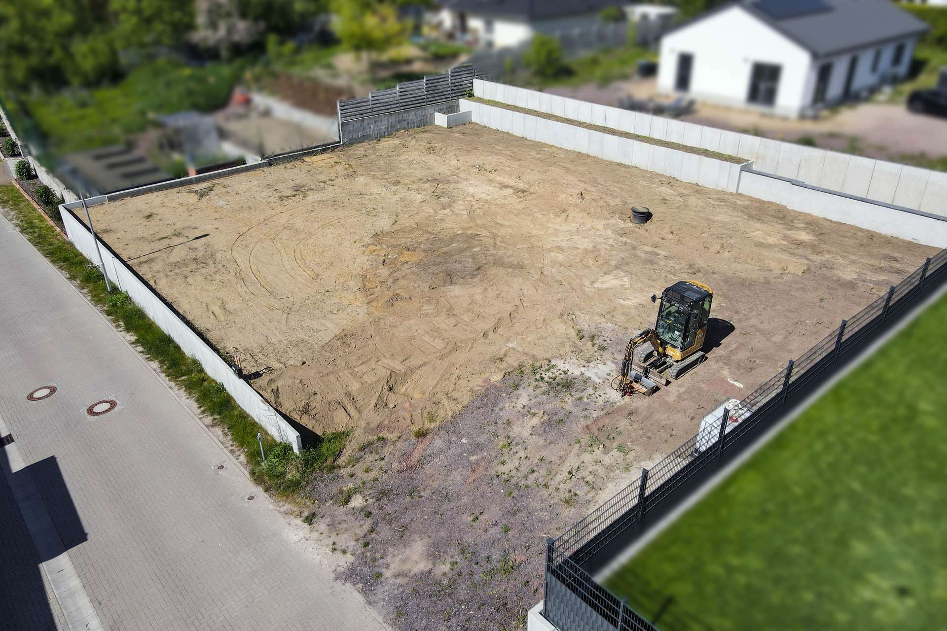 Leeres Baugrundstück mit einem Bagger, umgeben von einem Zaun und angrenzenden Straßen und Wohnhäusern.
