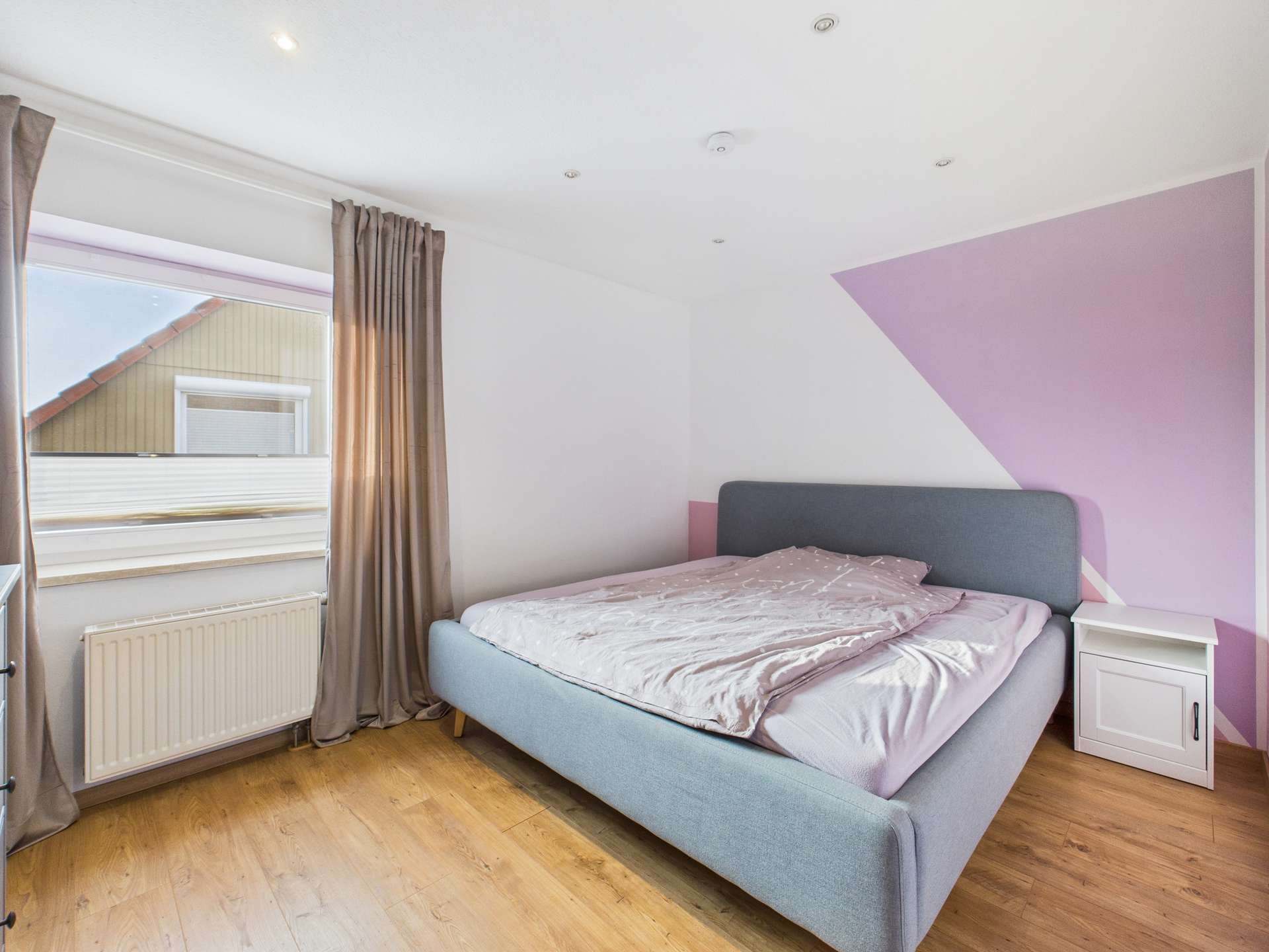 Schlafzimmer mit einem grauen Bett, weißen Wänden und einem Fenster mit Vorhängen. Holzfußboden und Nachttisch sichtbar.