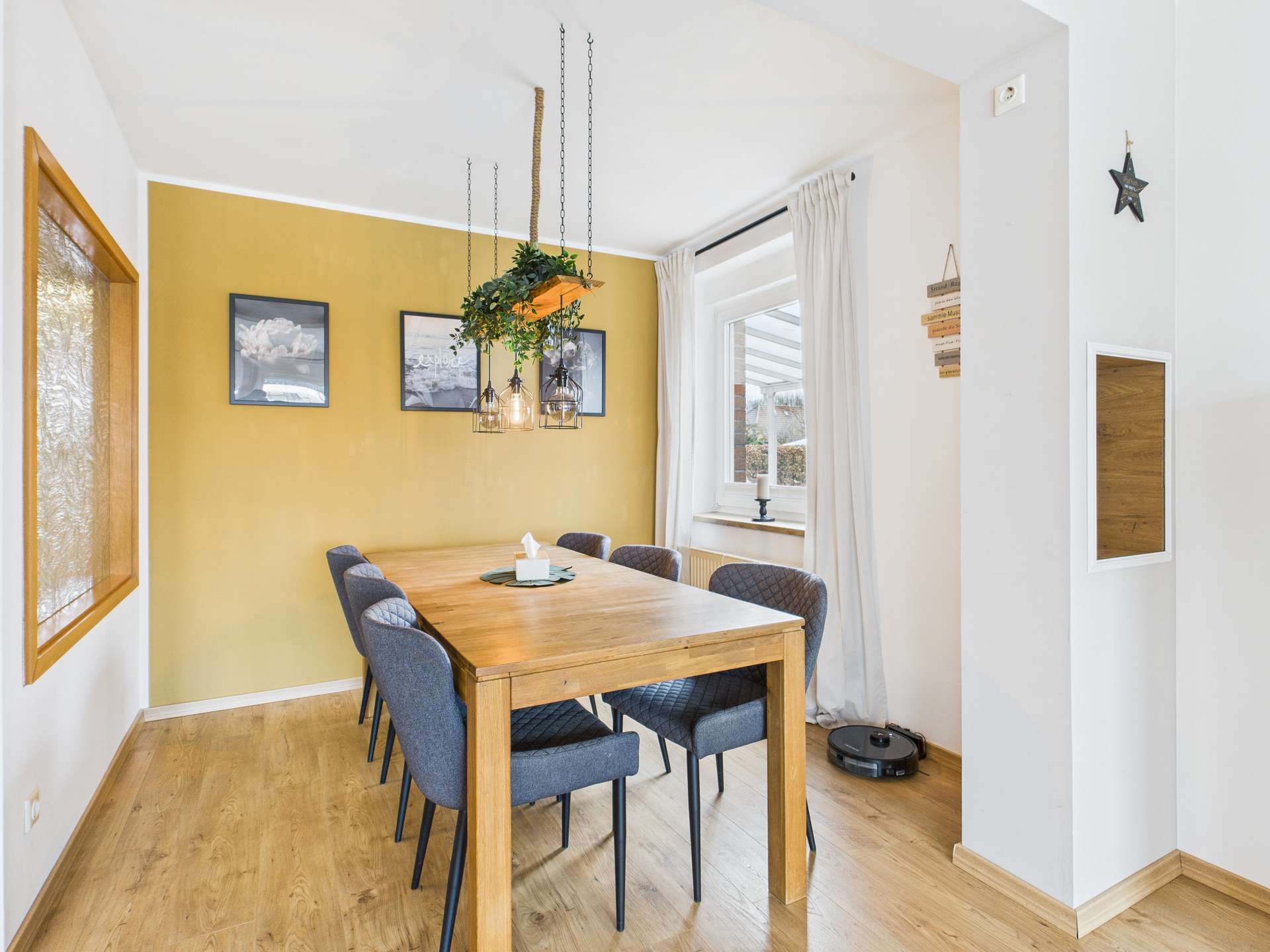 Esszimmer mit Holztisch, blauen Stühlen und einer goldenen Wand. Fenster mit Vorhängen und dekorativen Bildern.