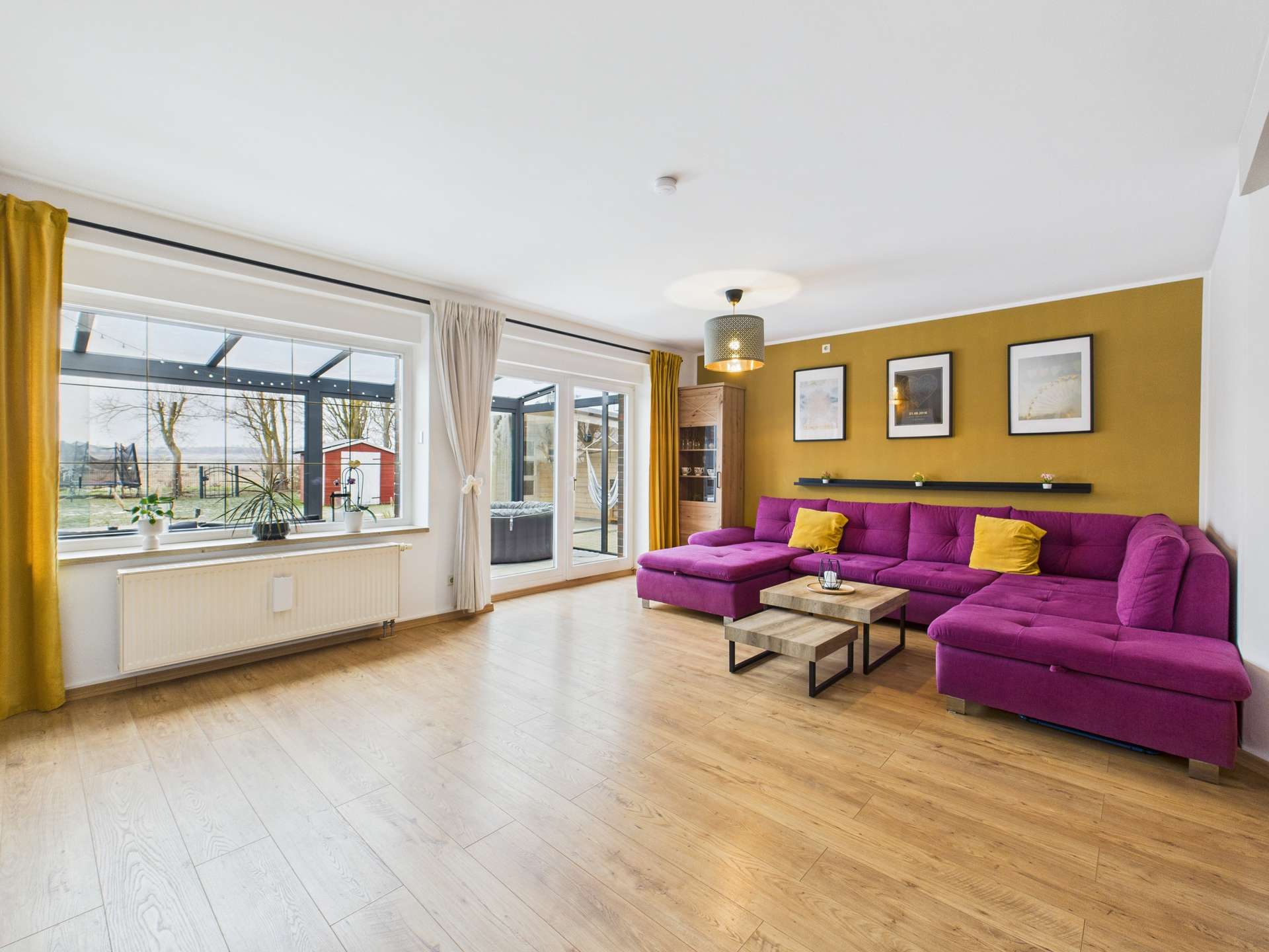 Wohnzimmer mit lila Sofa, Holztisch, großen Fenstern und gelben Vorhängen, Blick auf den Garten.