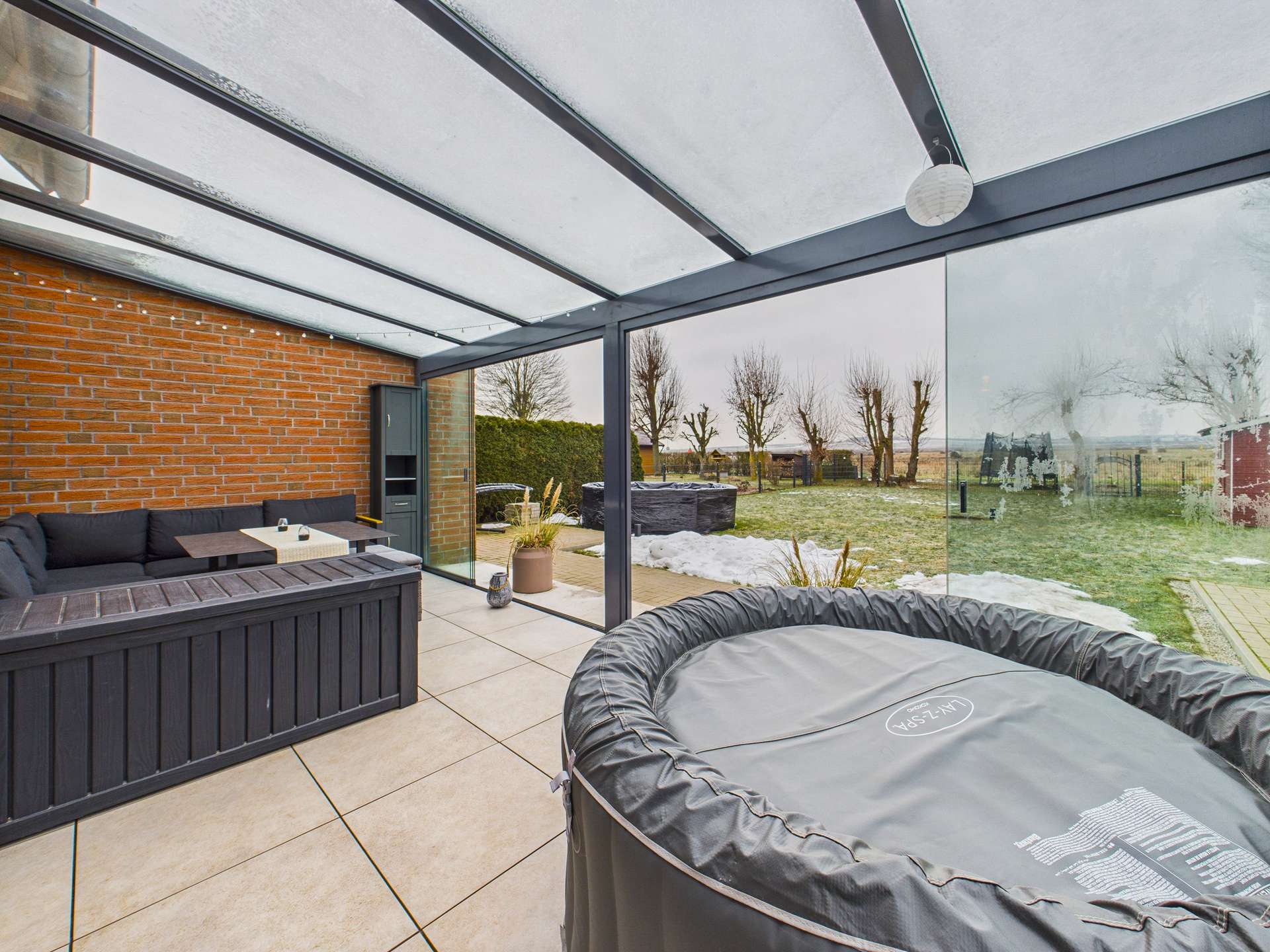 Überdachte Terrasse mit Sofa und einem runden Whirlpool, umgeben von einer grünen Wiese und Bäumen im Hintergrund.