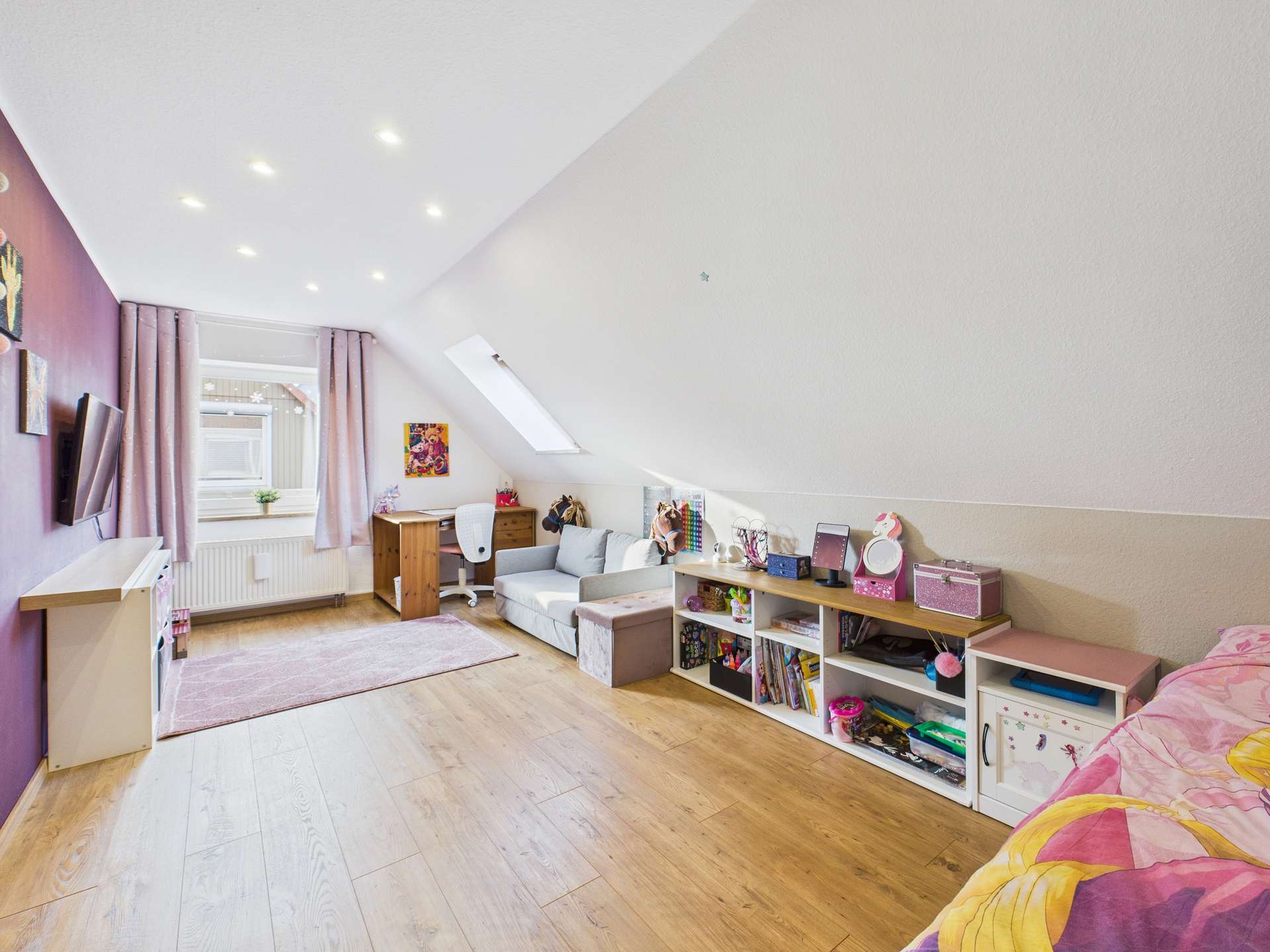 Ein helles, gemütliches Zimmer mit Dachschräge, Holzfußboden und Regalen voller Bücher und Spielzeug.