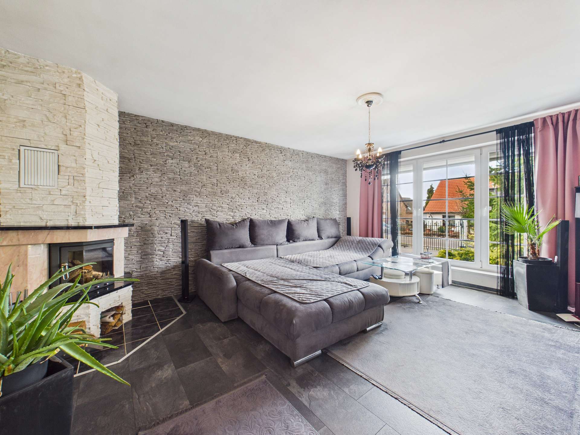 Modernes Wohnzimmer mit grauem Sofa, Kamin, Pflanzen und großen Fenstern, die auf einen Außenbereich blicken.
