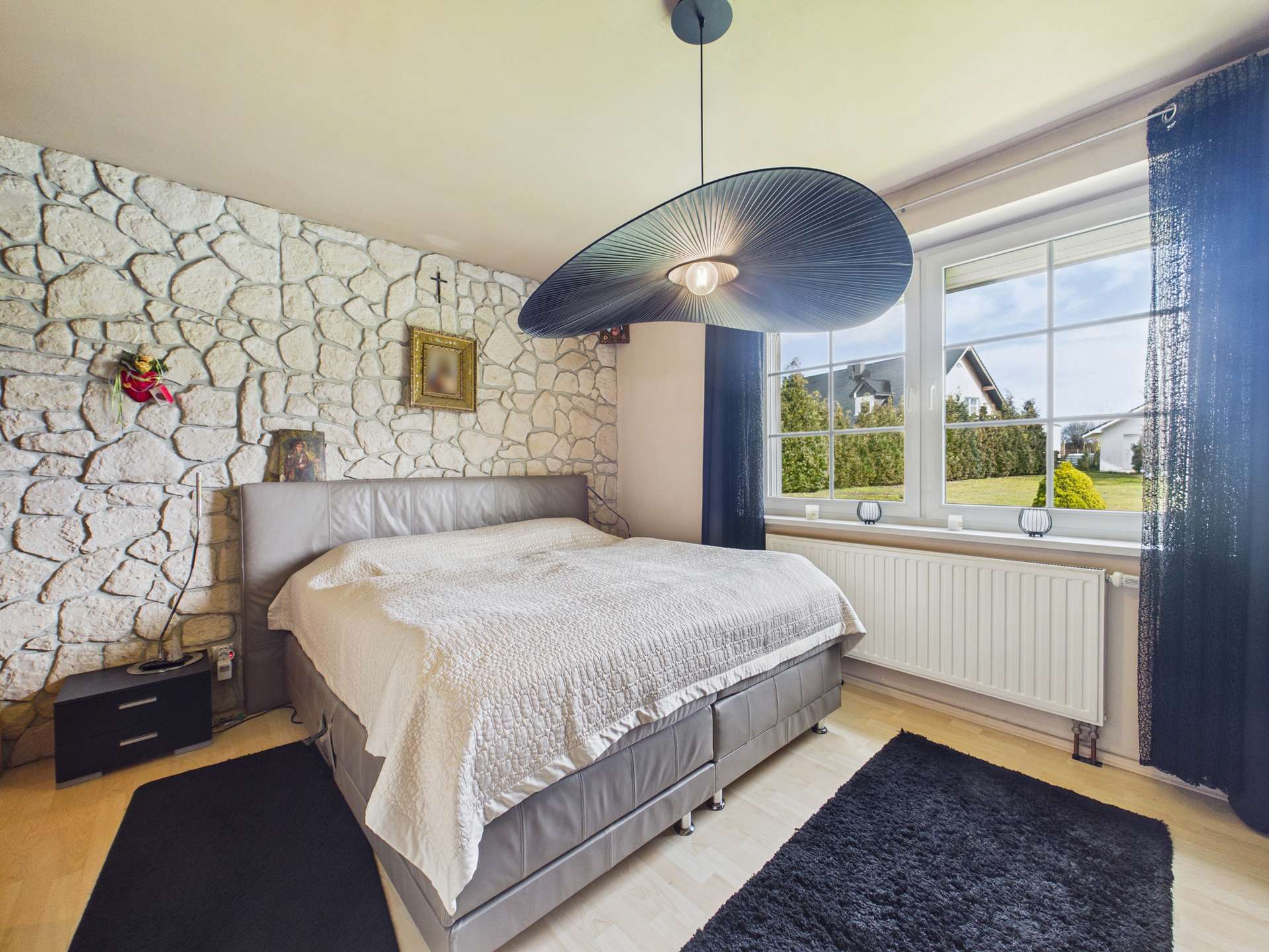 Helles Schlafzimmer mit einem großen Bett, Steinwand, modernem Lampendesign und Blick auf den Garten.