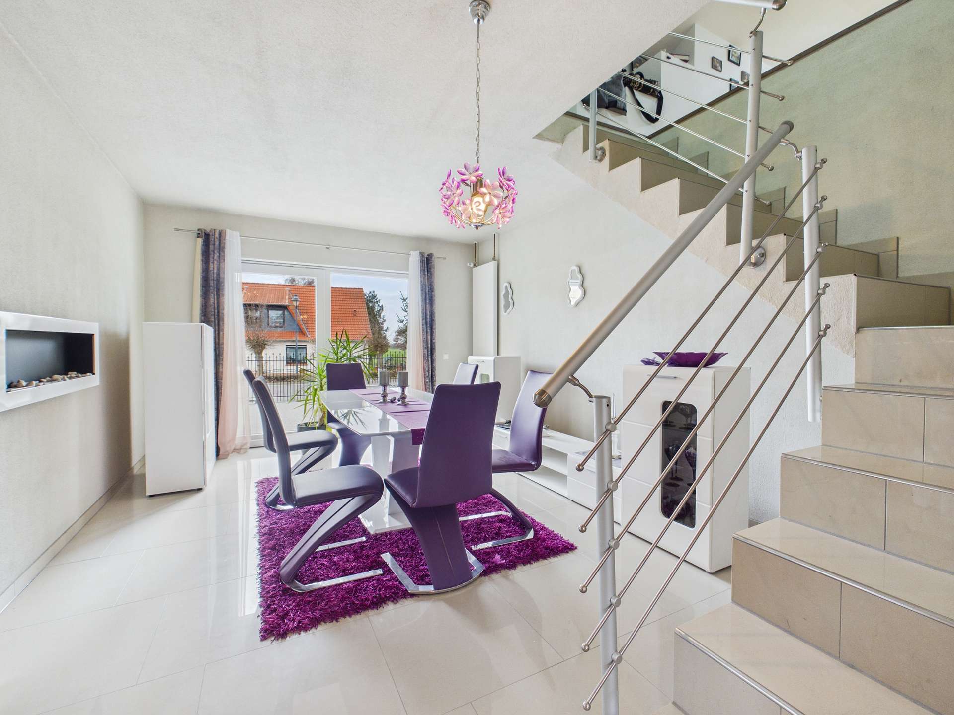 Helles Esszimmer mit lila Stühlen, Teppich und moderner Treppe. Großer Fensterblick auf den Garten.