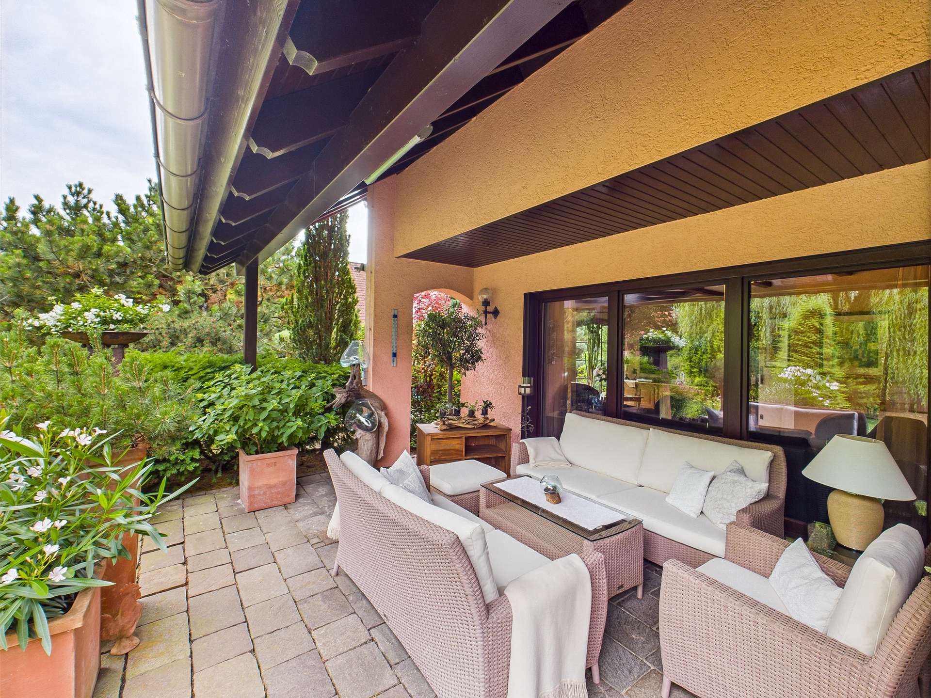 Gemütliche Terrasse mit einem Sofa, Tisch und Pflanzen, umgeben von Grün und einem modernen Haus.