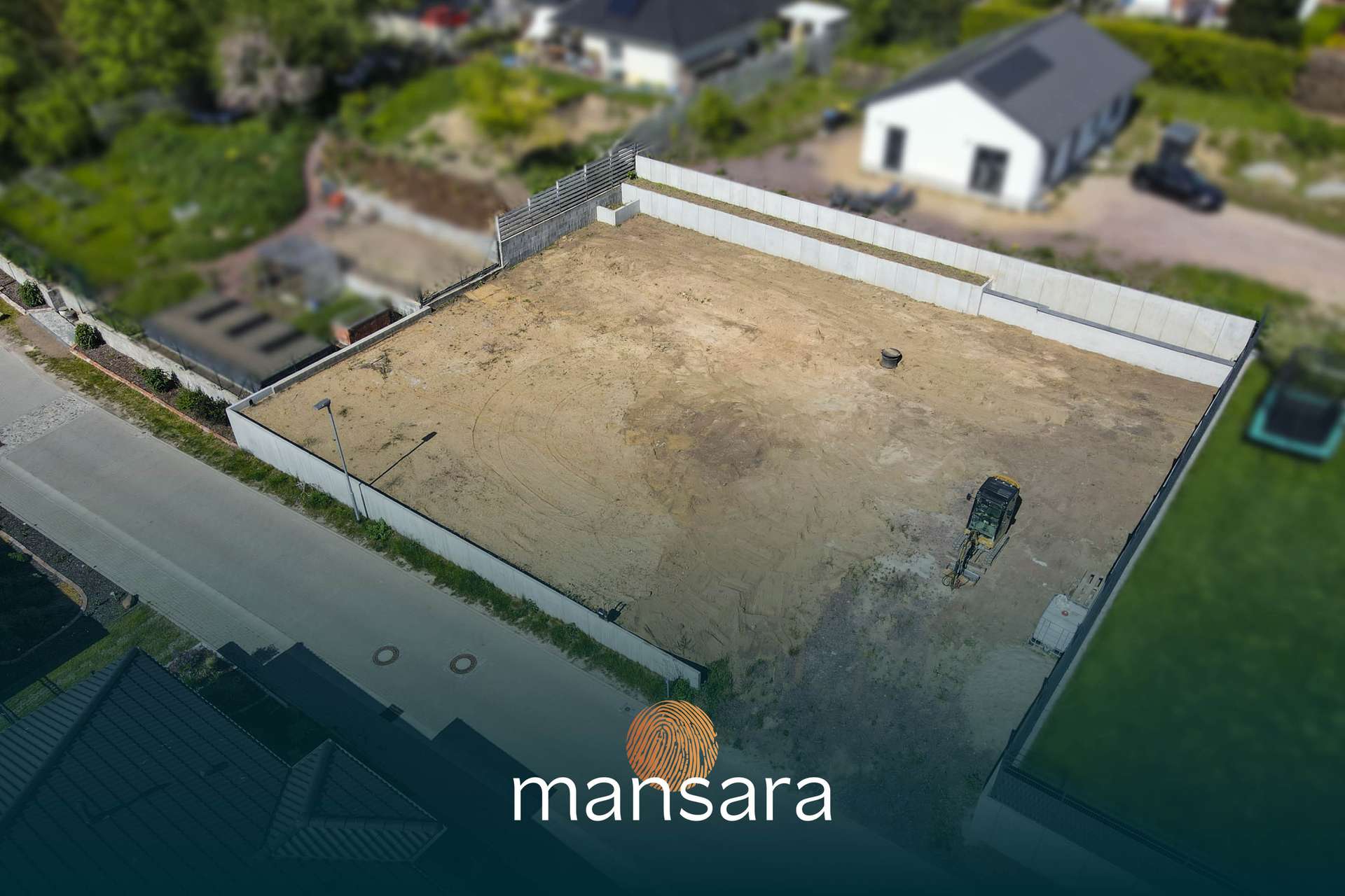 Leeres Grundstück mit Sandboden, umgeben von Mauern, in einer Wohngegend mit einigen Häusern im Hintergrund.