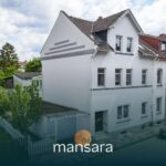 Moderne Wohnhausfassade mit grauen Elementen, umgeben von Grünflächen und einem Parkplatz.