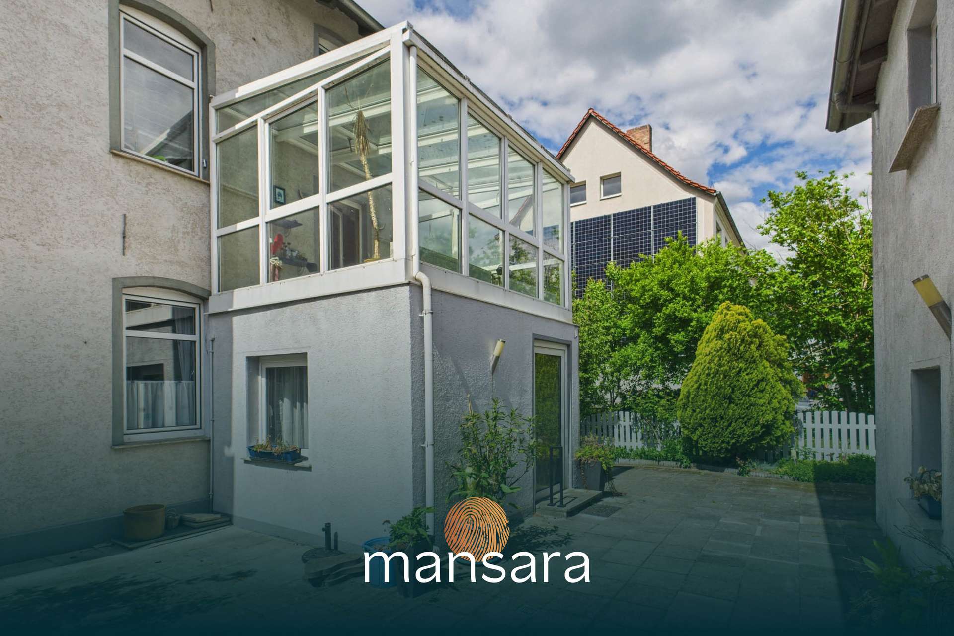 Glasveranda an einem Wohnhaus mit Garten und angrenzenden GebÀuden, unter einem bewölkten Himmel.